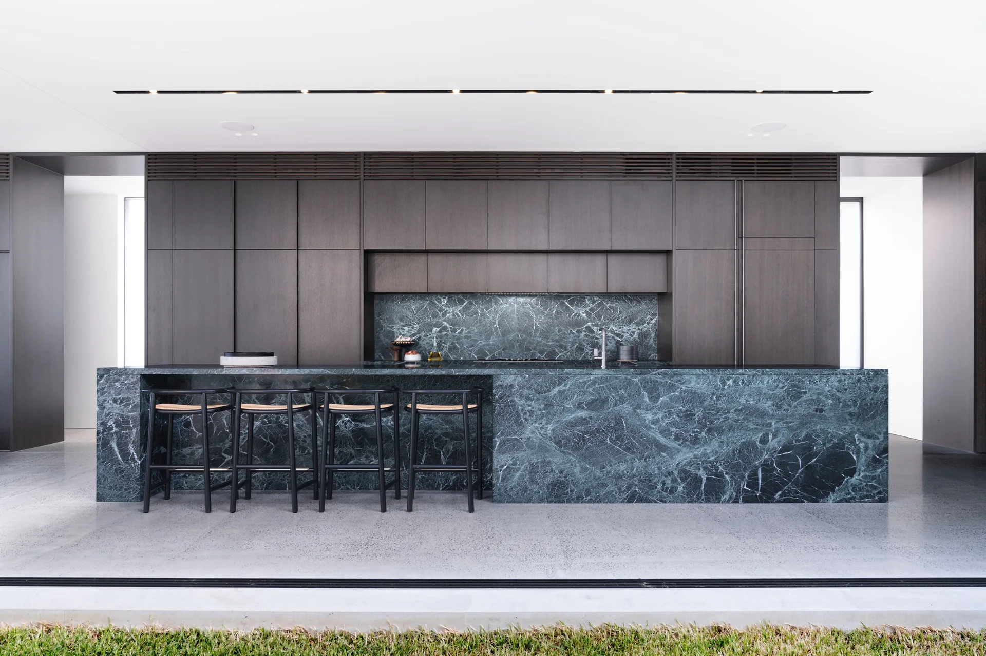 A modern kitchen featuring a large green marble island with black bar stools, matching marble backsplash, and full-height dark wood cabinetry.