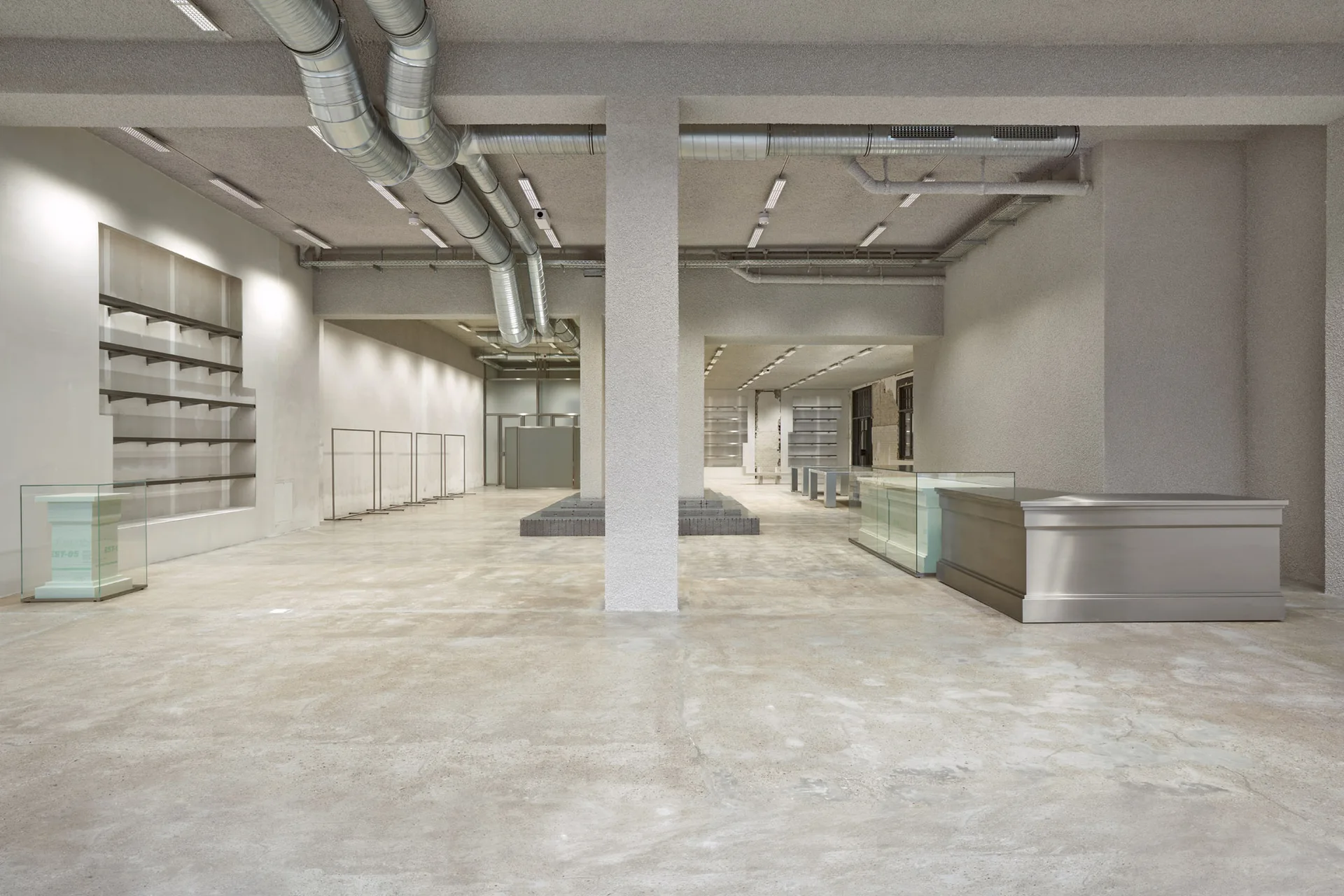 A large minimalist retail space features a polished concrete floor, exposed silver overhead ducting, textured gray walls, and a metallic display counter.