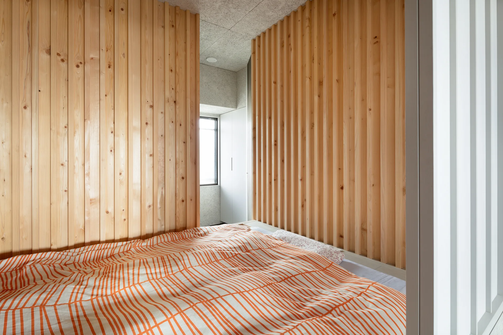 A bed with a striped orange and white duvet is nestled between two vertical wood slat partition walls under a textured concrete ceiling.