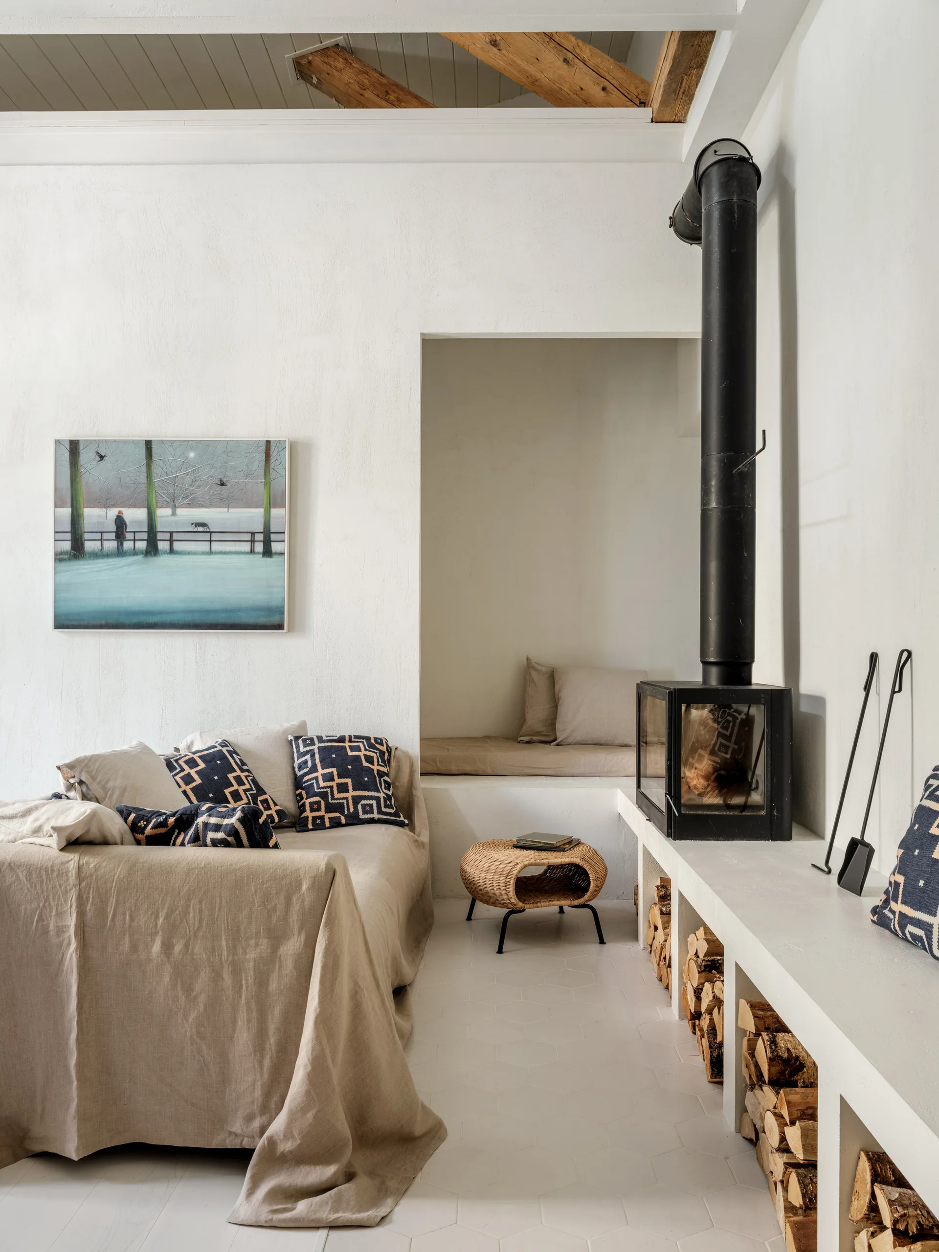 A minimalist living room featuring a linen-covered sofa, a black wood stove on a plastered hearth with firewood storage, and a recessed seating nook.