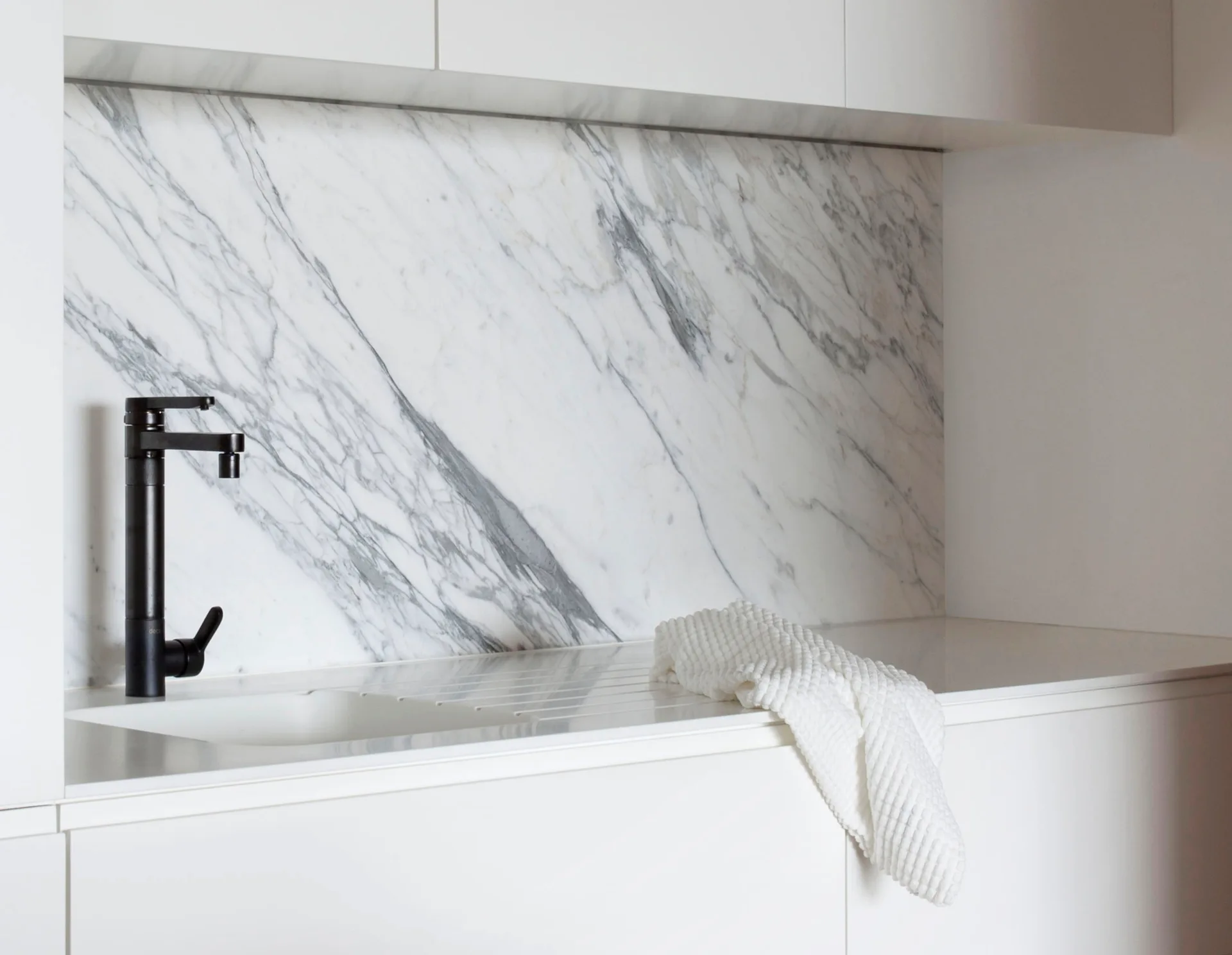 A minimalist white kitchen counter features an integrated sink with a black faucet, set against a heavily veined grey and white marble backsplash.