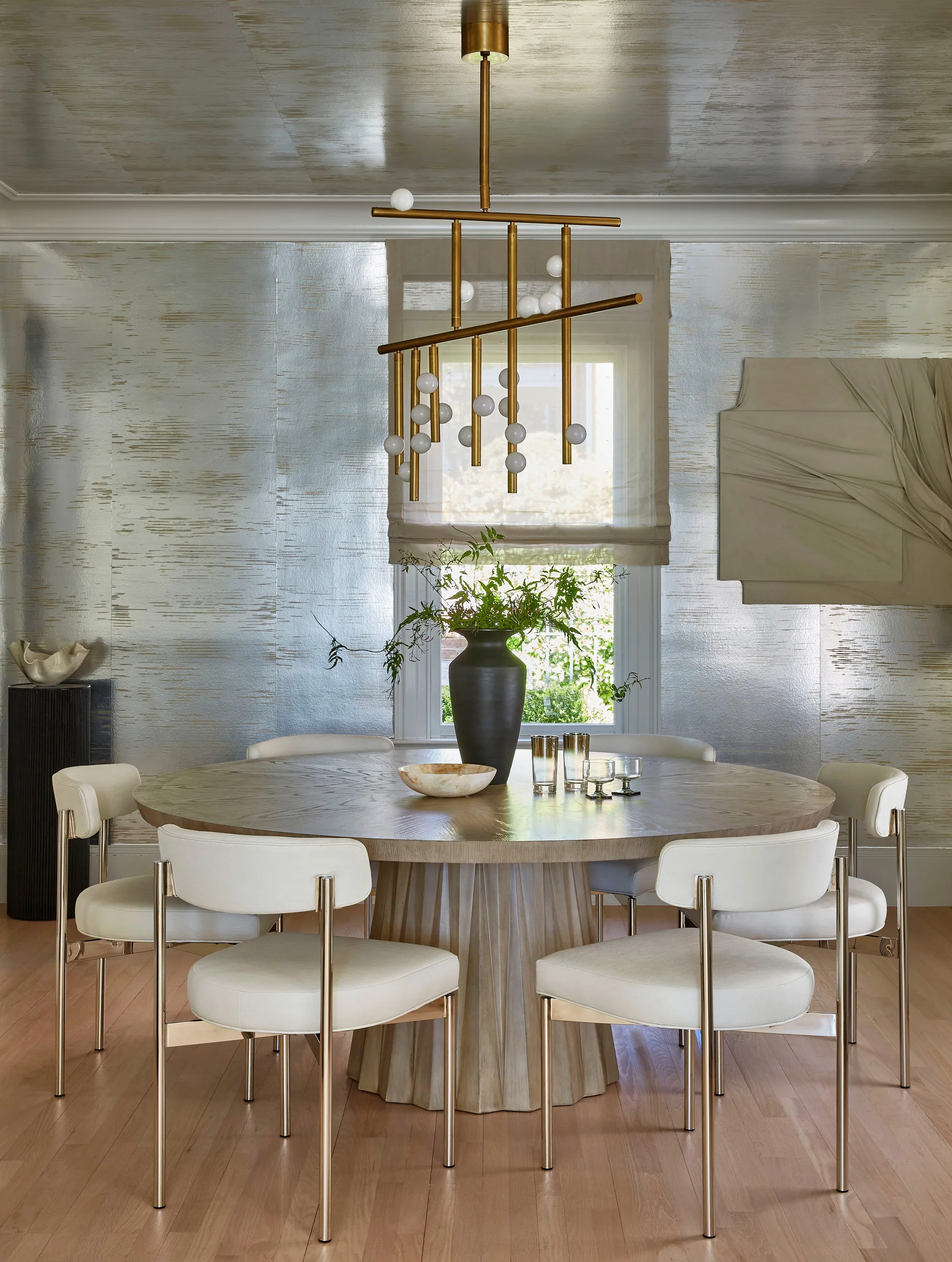 A round wooden dining table surrounded by white chairs sits beneath a gold tiered chandelier in a room with silver textured wallpaper.