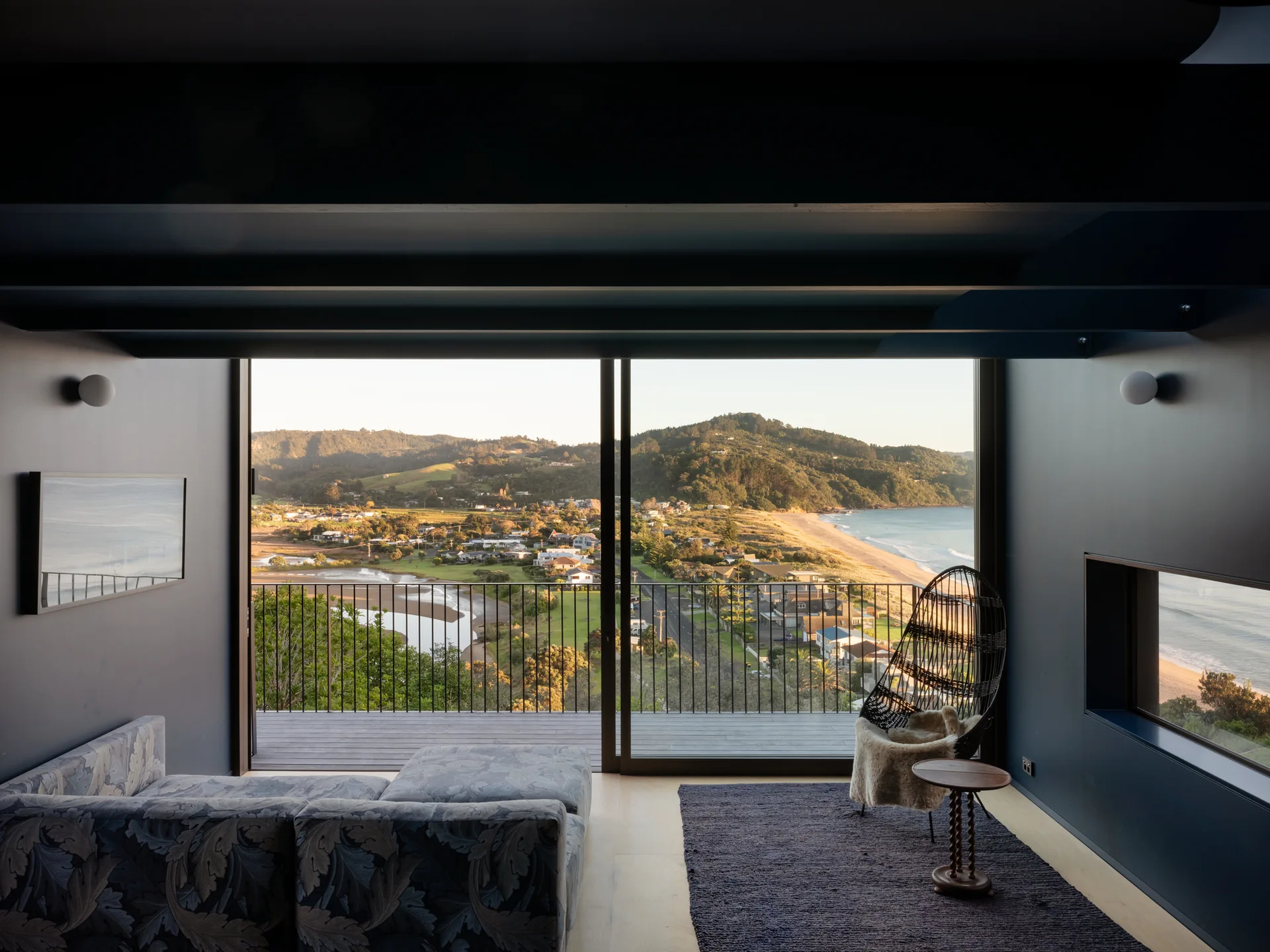 A modern navy blue living room features a patterned sofa, a rattan lounge chair, and large sliding glass doors opening to a scenic coastal balcony.