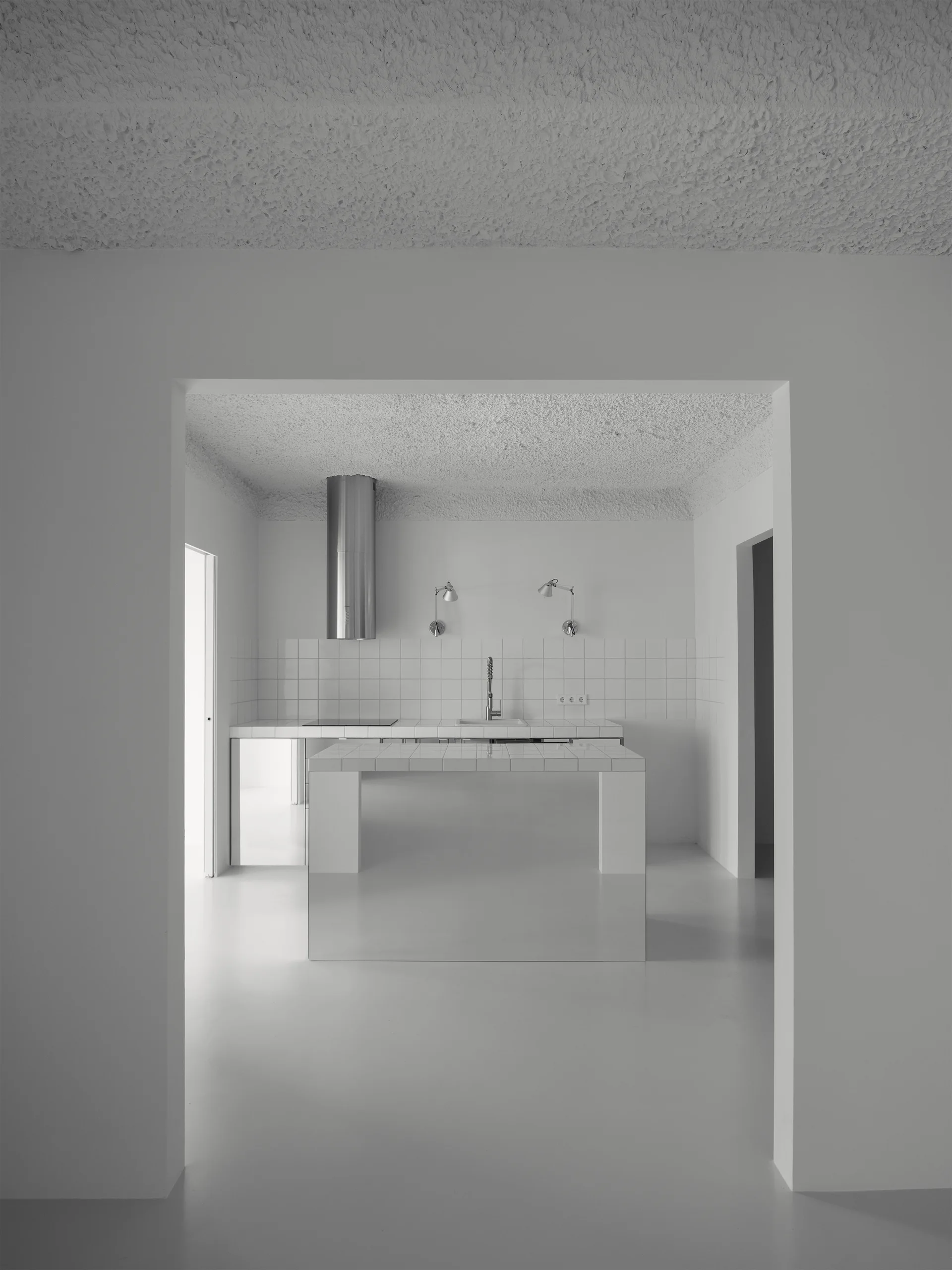 A minimalist white kitchen framed by an entryway, featuring a square-tiled kitchen island, matching tiled countertops, and a cylindrical steel range hood.