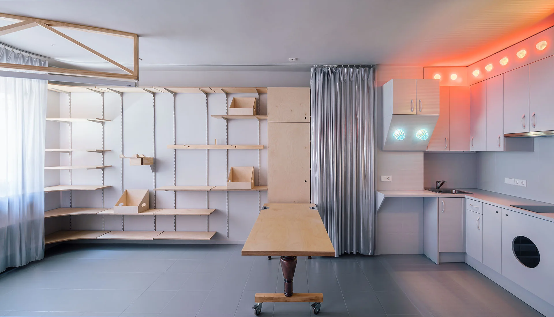 A minimalist kitchen space featuring adjustable wood shelving, a mobile wooden island table on wheels, and white cabinets next to a silver curtain.