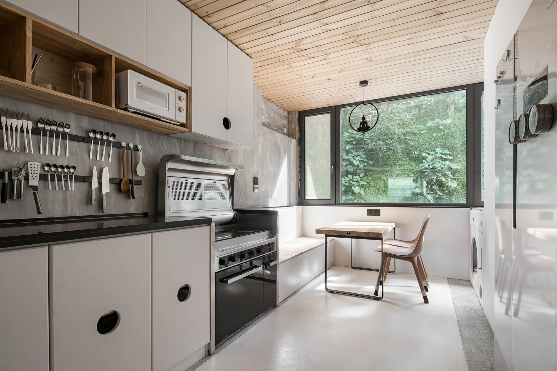 Modern kitchen featuring white cabinetry, raw concrete walls, a slatted wood ceiling, and a small dining nook with a built-in bench by a window.