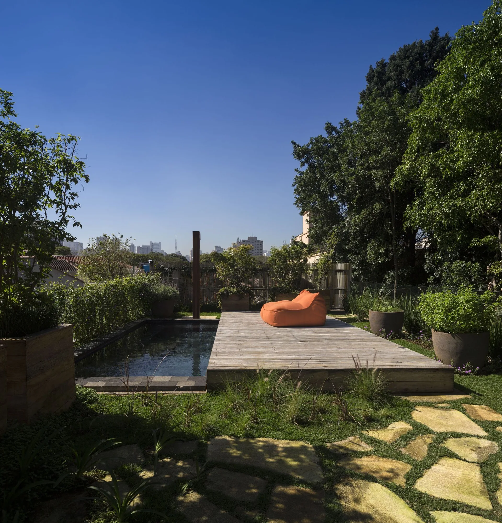 An outdoor rectangular swimming pool features an adjacent weathered wooden deck with an orange bean bag lounge chair, surrounded by a lush lawn and trees.