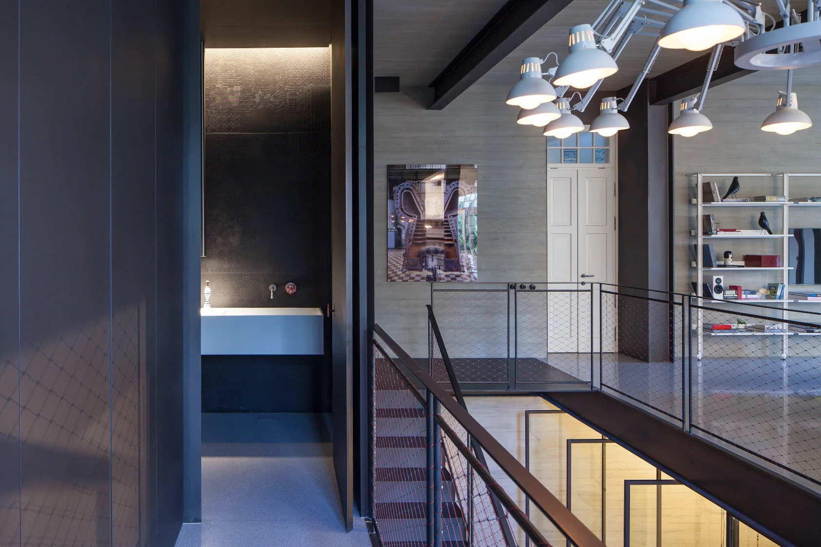 A mezzanine landing featuring exposed concrete walls, a dark metal staircase railing with red mesh, and a white multi-light chandelier.