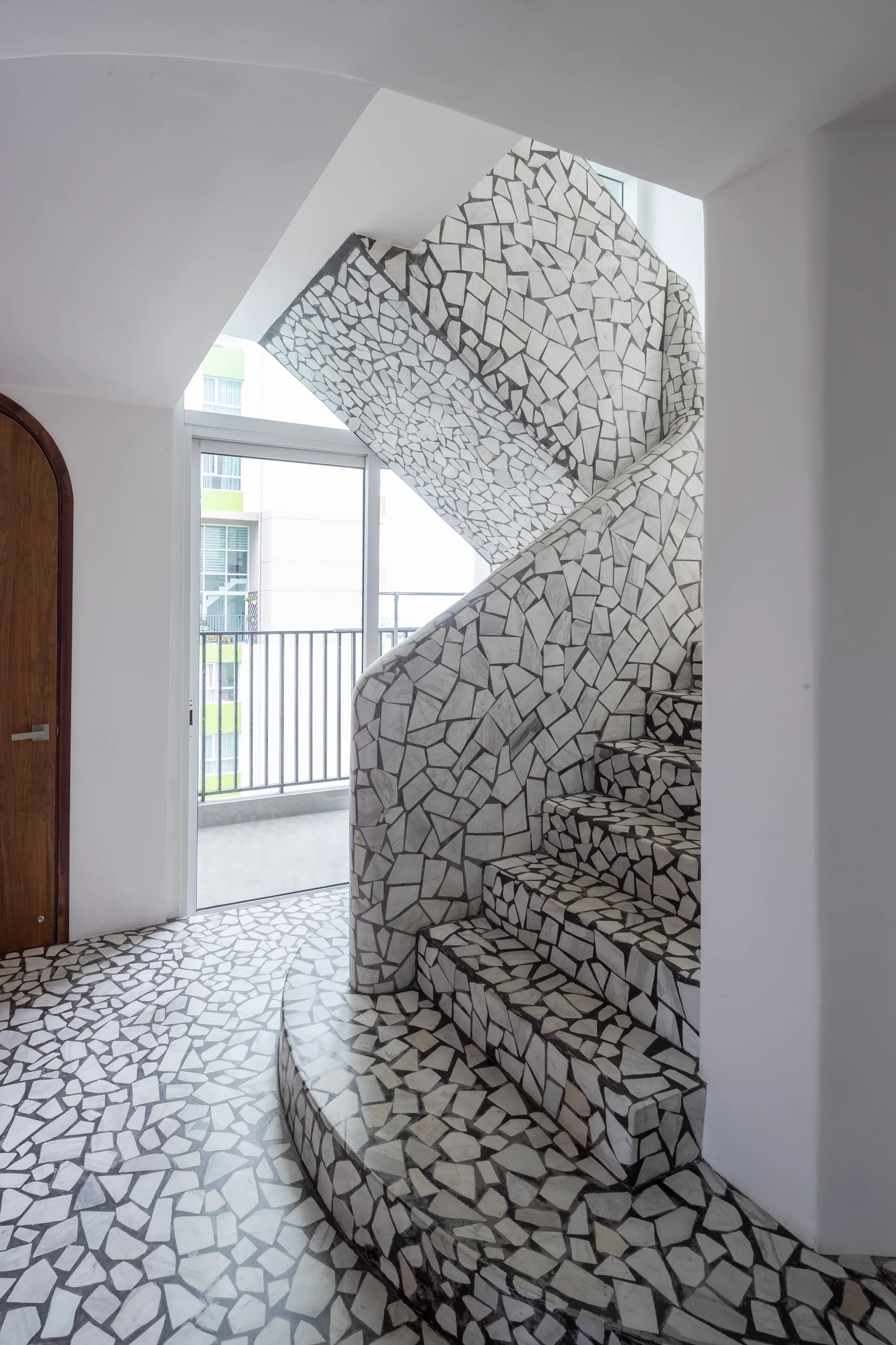 A curved indoor staircase and entryway floor covered in white and grey irregular stone mosaic tiles, next to plain white walls and an arched wooden door.