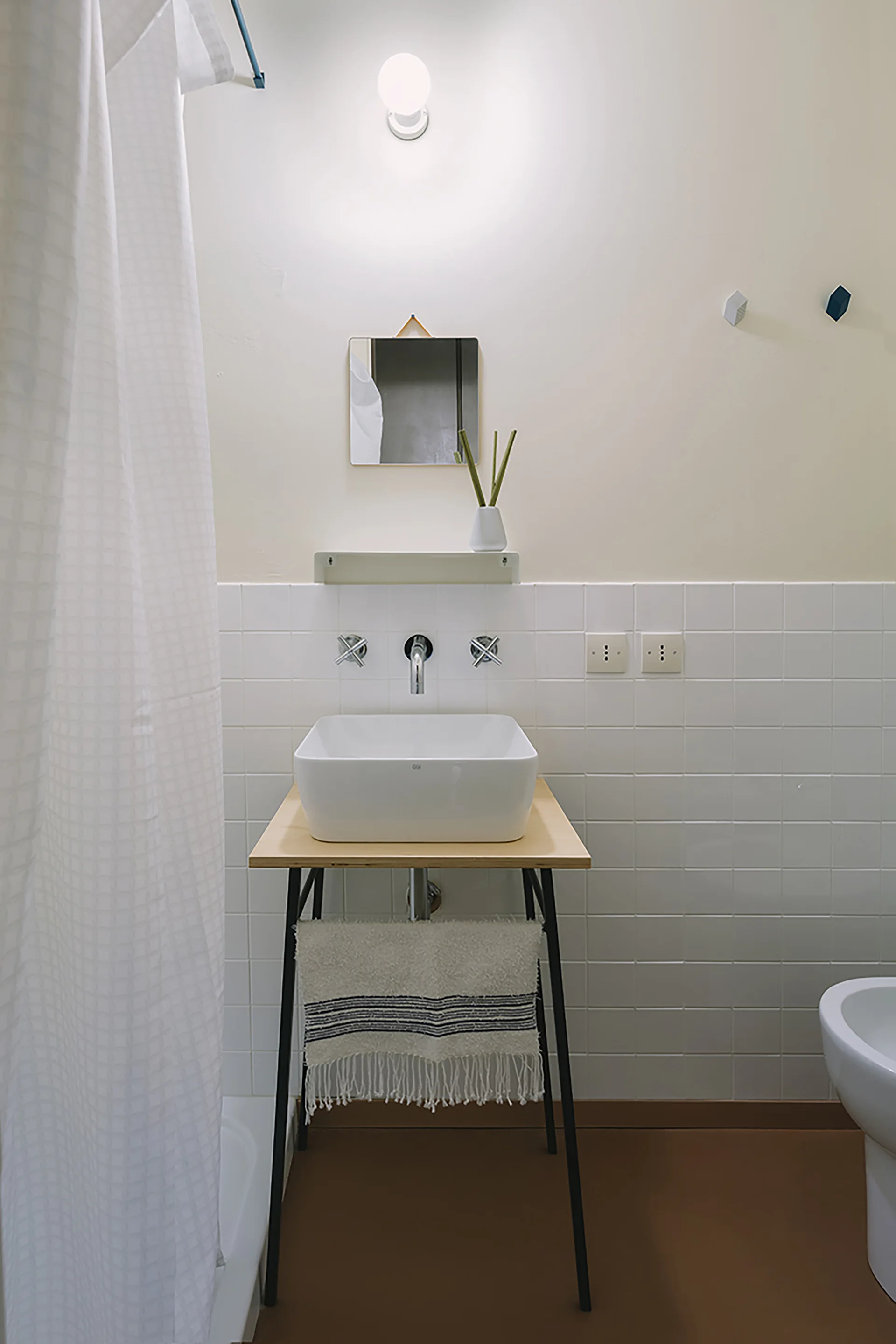 A minimalist bathroom features a white vessel sink on a wood-topped vanity stand against a white square-tiled wall.