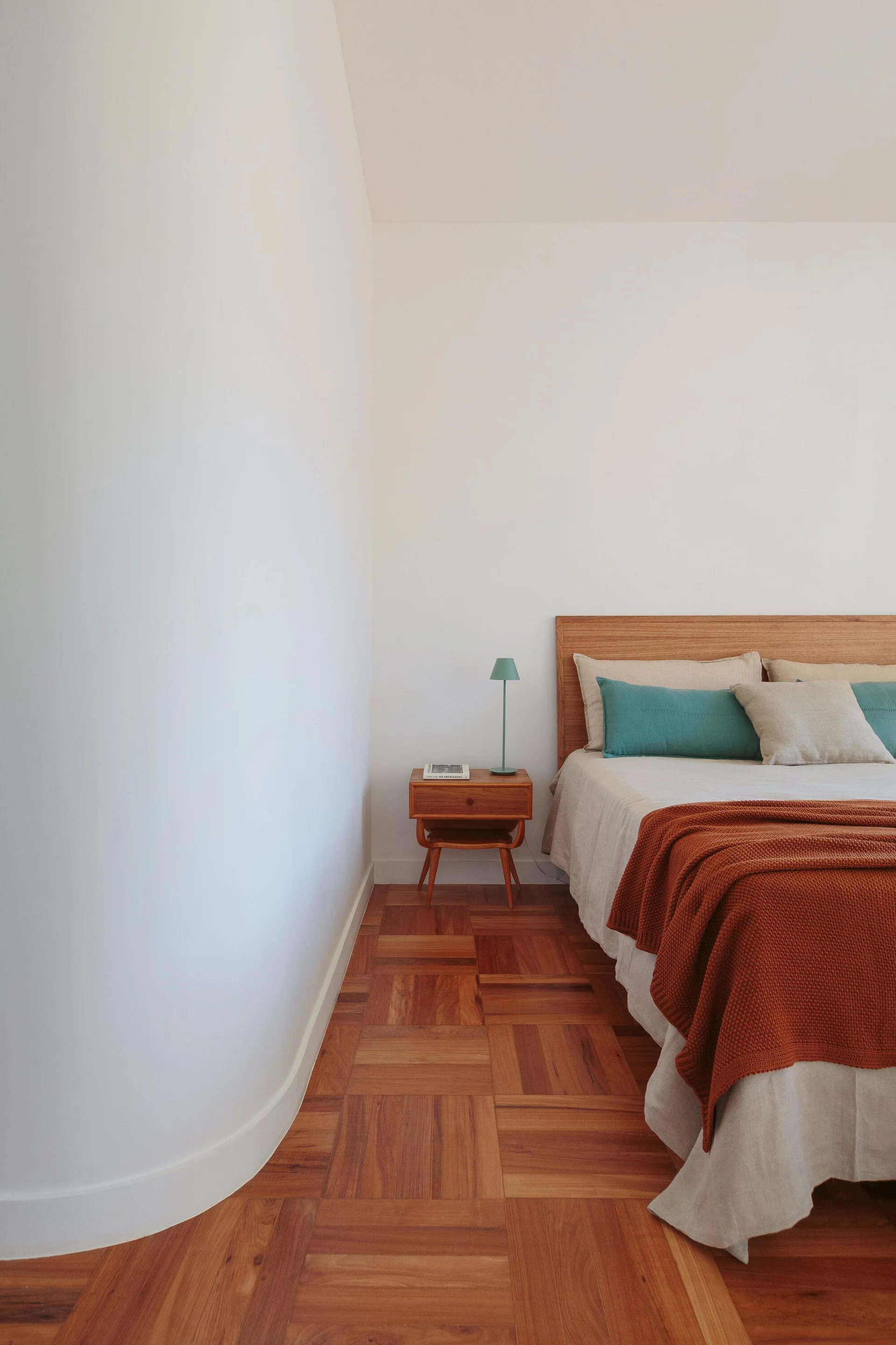A minimalist bedroom featuring a wooden bed with a terracotta throw blanket, a mid-century bedside table, and patterned parquet wood flooring.