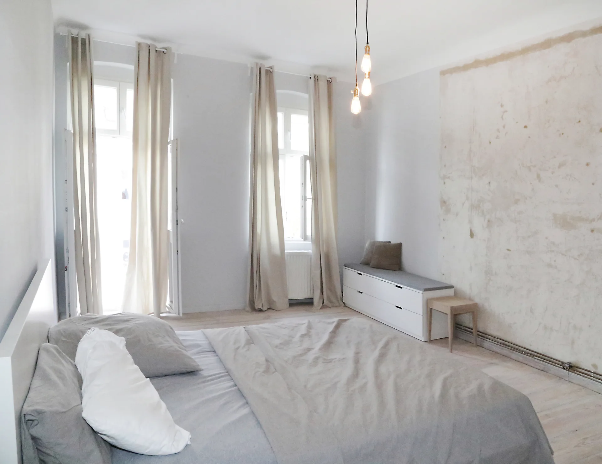 A minimalist bedroom features a bed with grey linens, two tall windows with beige curtains, and a textured unfinished plaster wall on the right.
