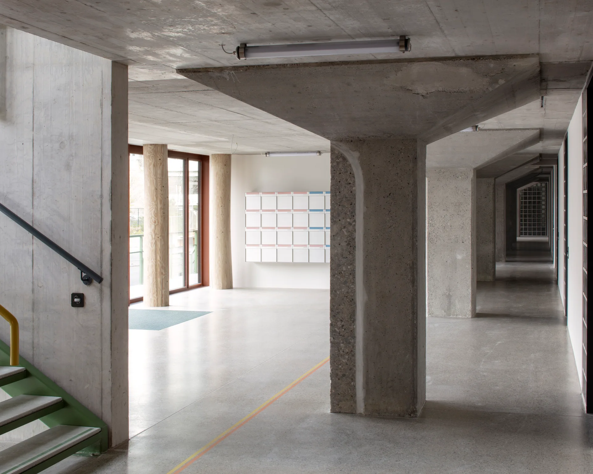 A minimalist lobby features large exposed concrete columns, round wooden pillars, and a grid of white mailboxes on a far wall.
