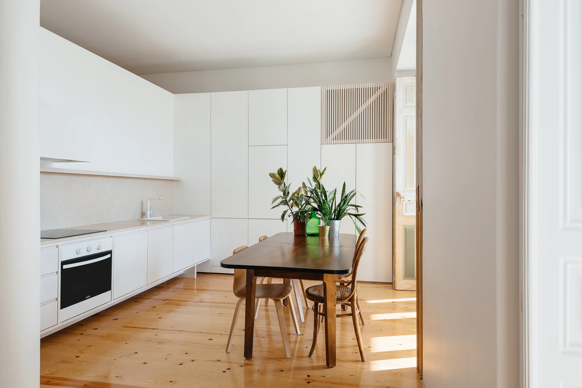 A minimalist kitchen features white flat-front cabinetry, a dark wood dining table with potted plants, and light-toned pine flooring.