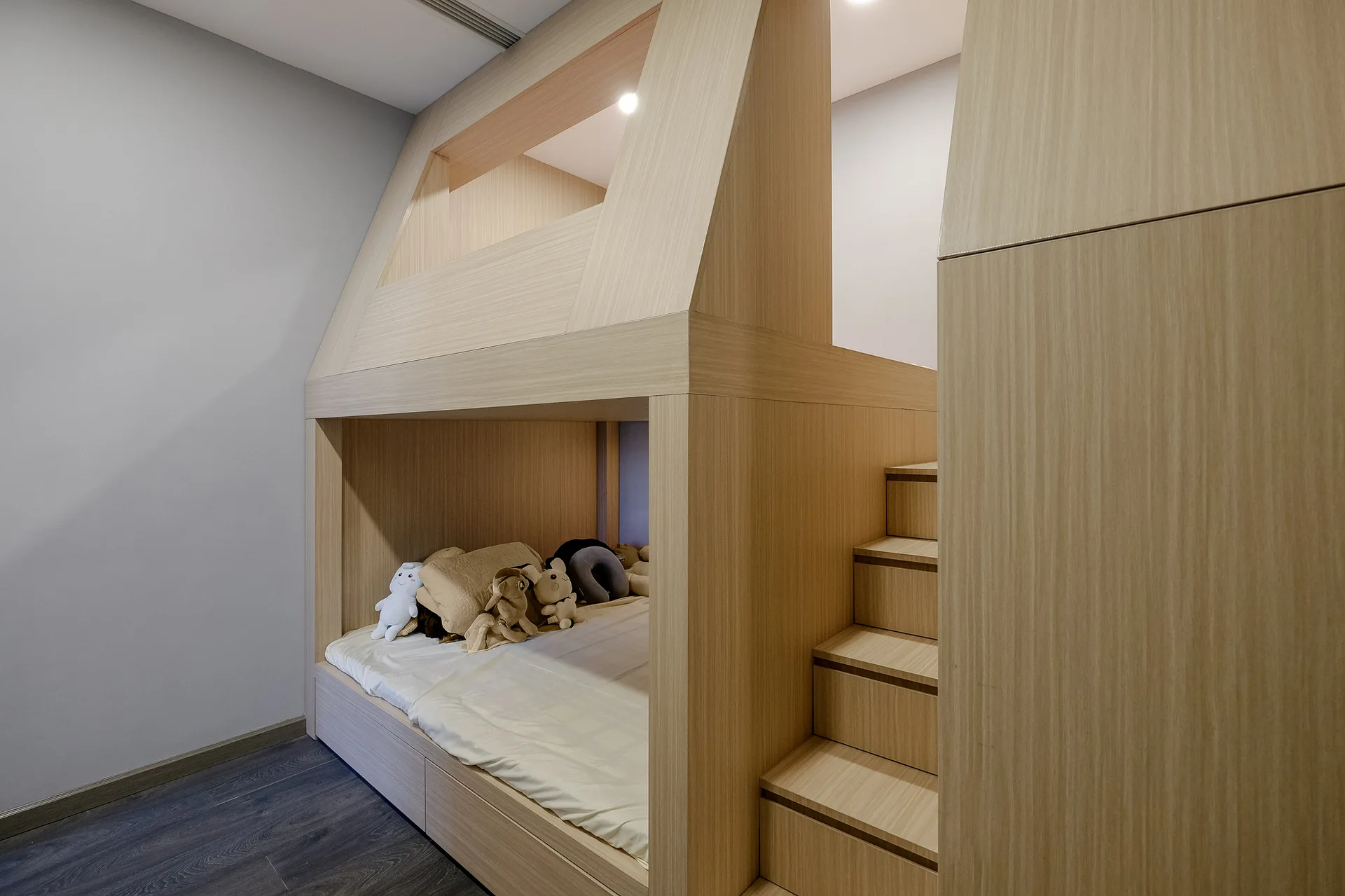 A custom light wood bunk bed with built-in stairs, a lower mattress with stuffed animals, and a lofted upper level.