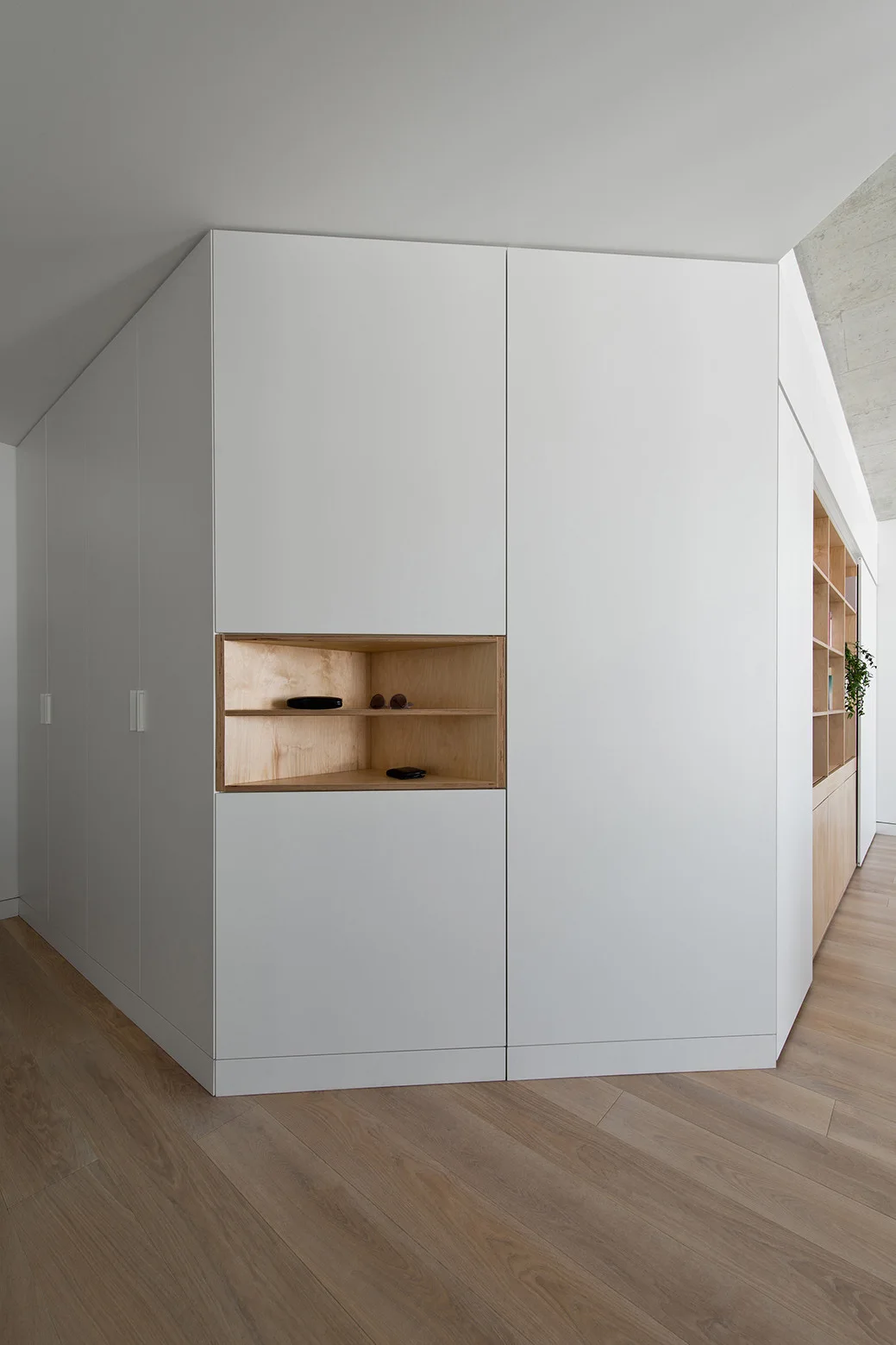 A floor-to-ceiling white minimalist storage unit features a recessed light wood display shelf set into the corner above light oak wood floors.
