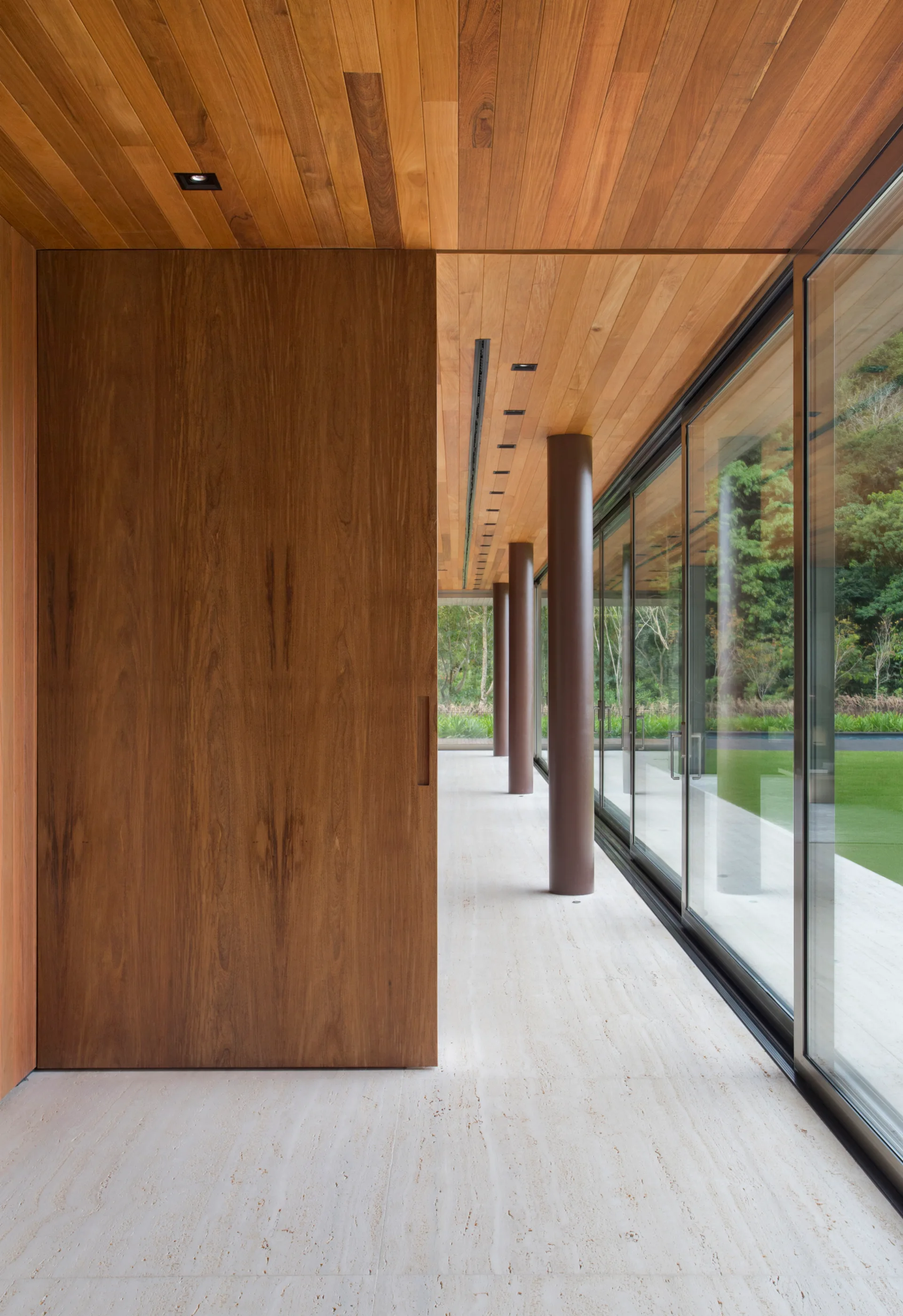 A modern hallway features a wood plank ceiling, a large sliding wood door, travertine stone flooring, and floor-to-ceiling glass walls.