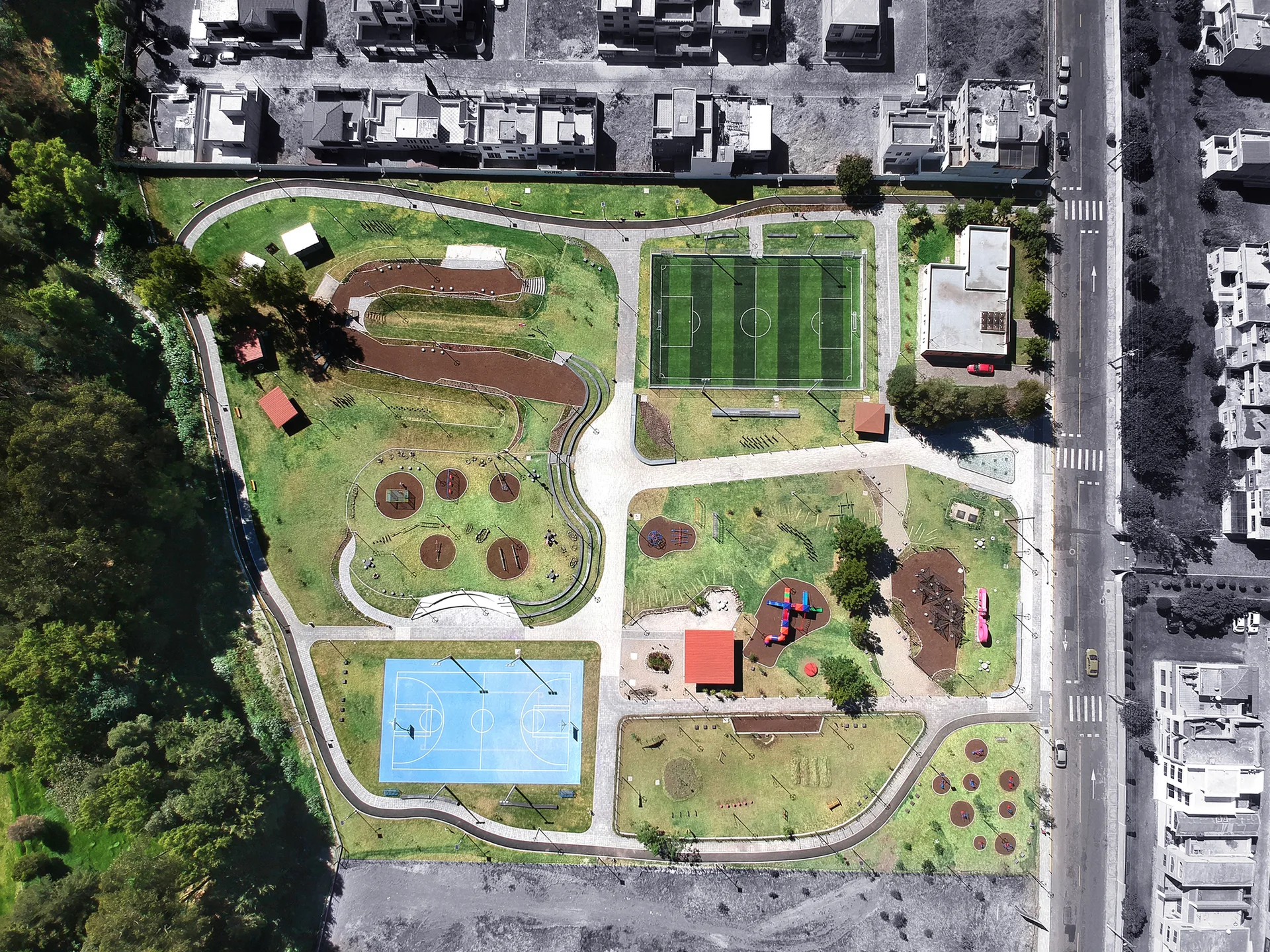 An aerial view of a landscaped public park featuring a green soccer field, a blue basketball court, and winding paved pathways.