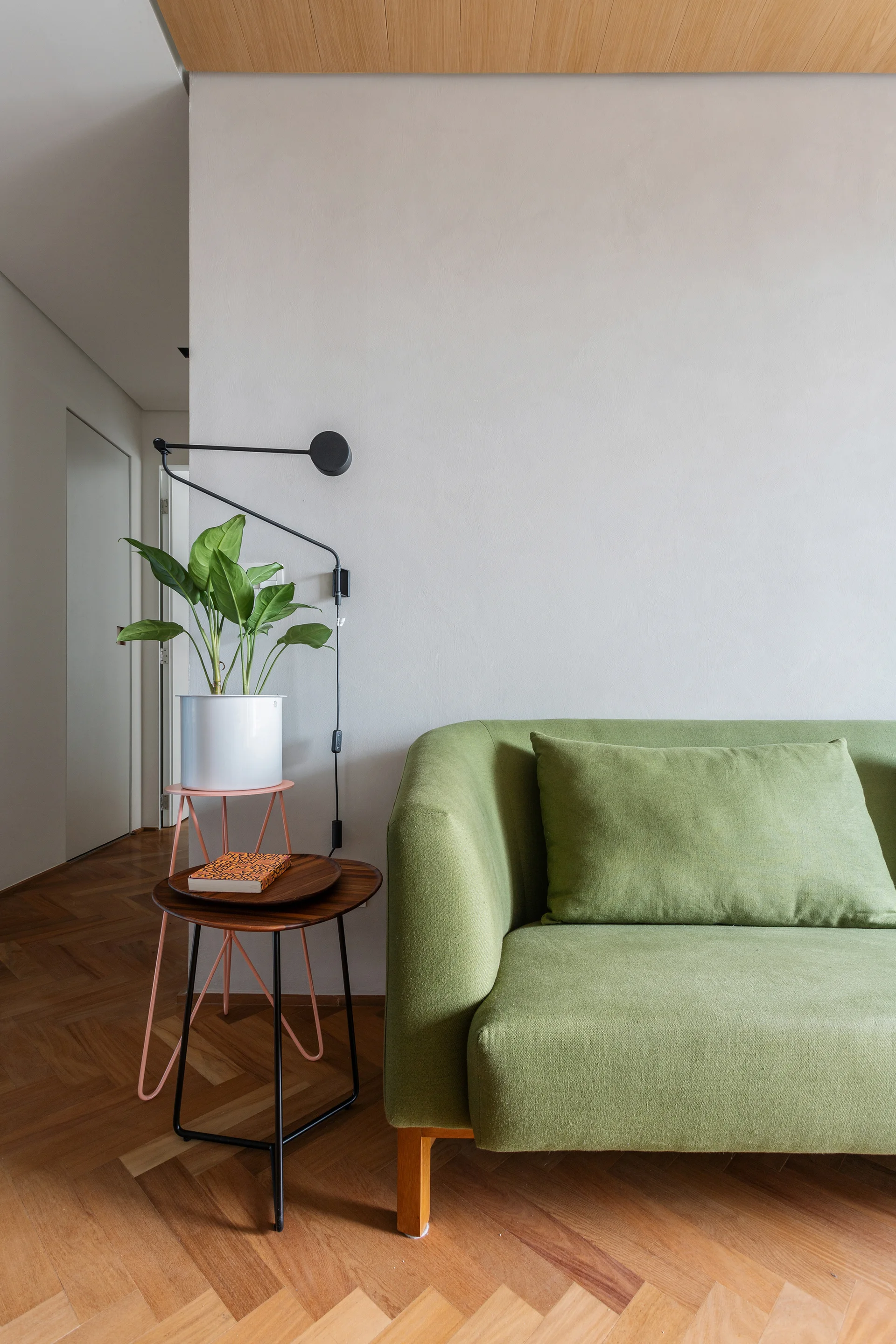A green upholstered sofa sits on herringbone wood flooring next to nested side tables, a potted plant, and a black adjustable wall lamp.