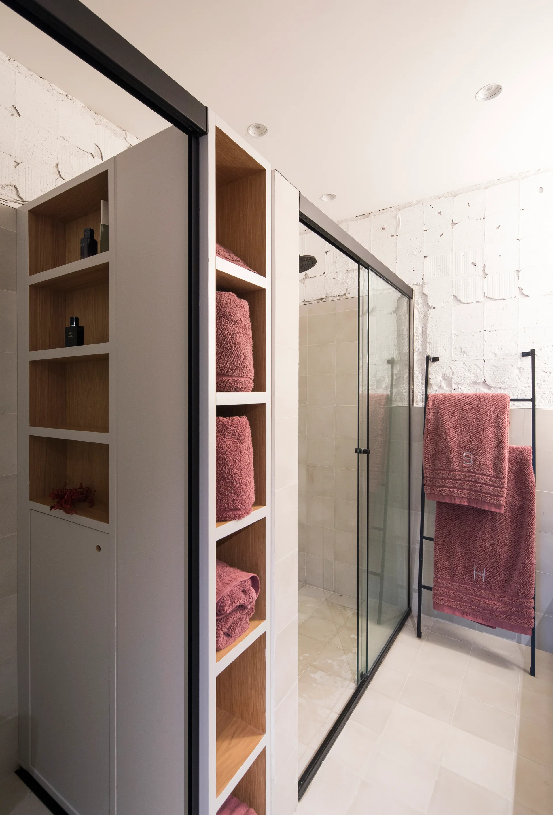 A modern bathroom features a tall white and wood shelving unit holding mauve towels next to a glass-enclosed shower stall and black ladder rack.