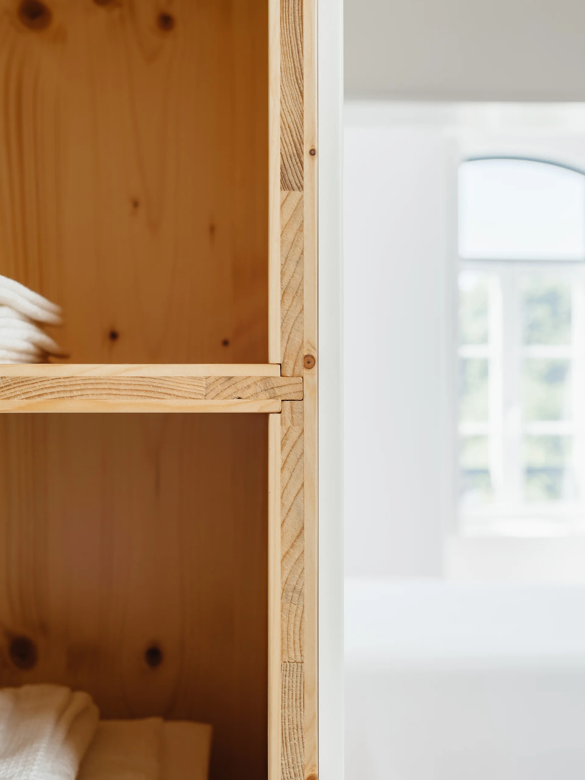 A close-up view of a natural wood closet shelving unit with exposed joinery and stacked white towels, set against a bright, soft-focus background.