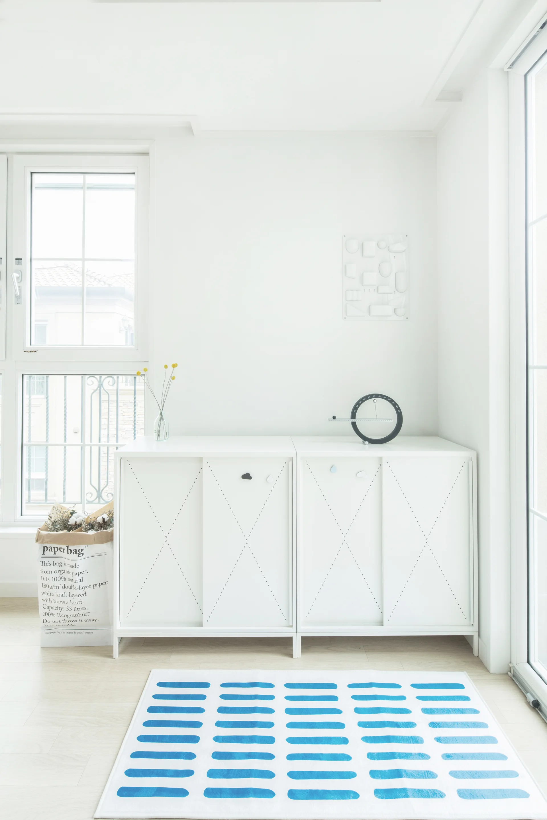 A white minimalist console cabinet with patterned sliding doors sits on light wood flooring next to a blue striped rug and large windows.