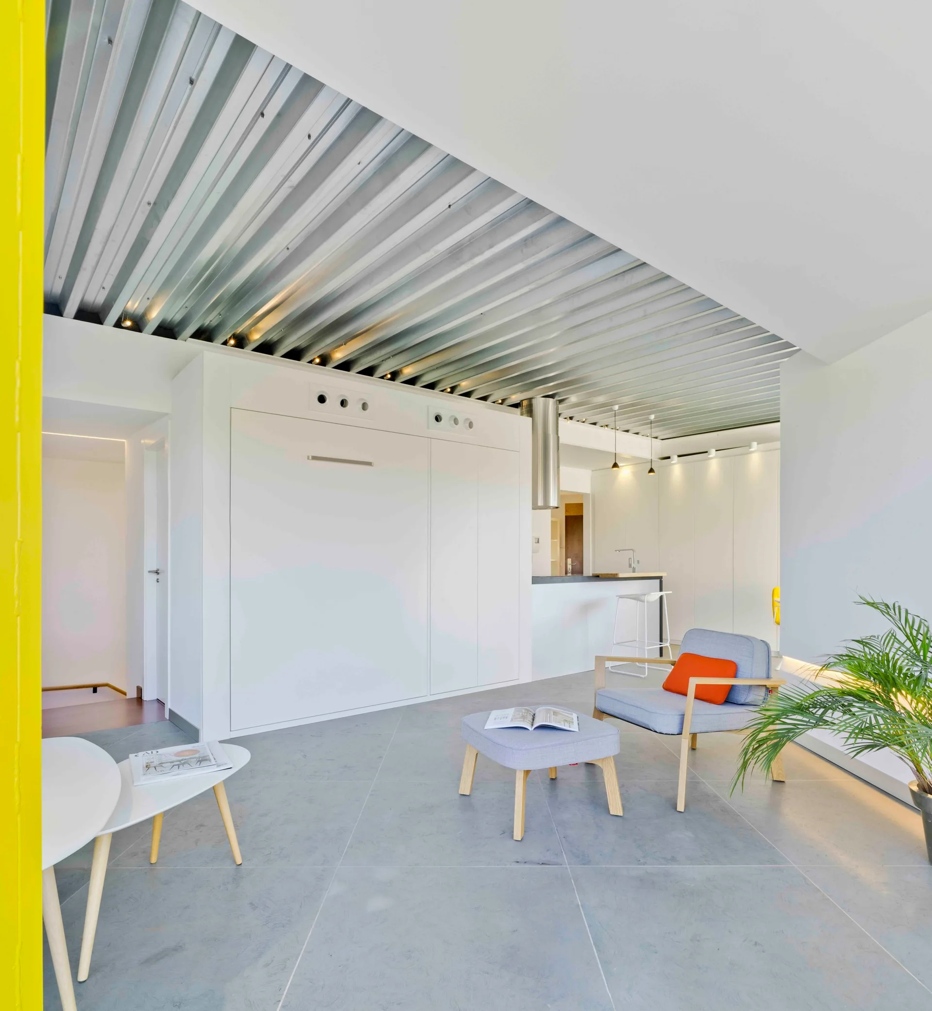 A minimalist living space featuring a corrugated metal ceiling, integrated white wall cabinets, and a light blue armchair with an orange cushion.
