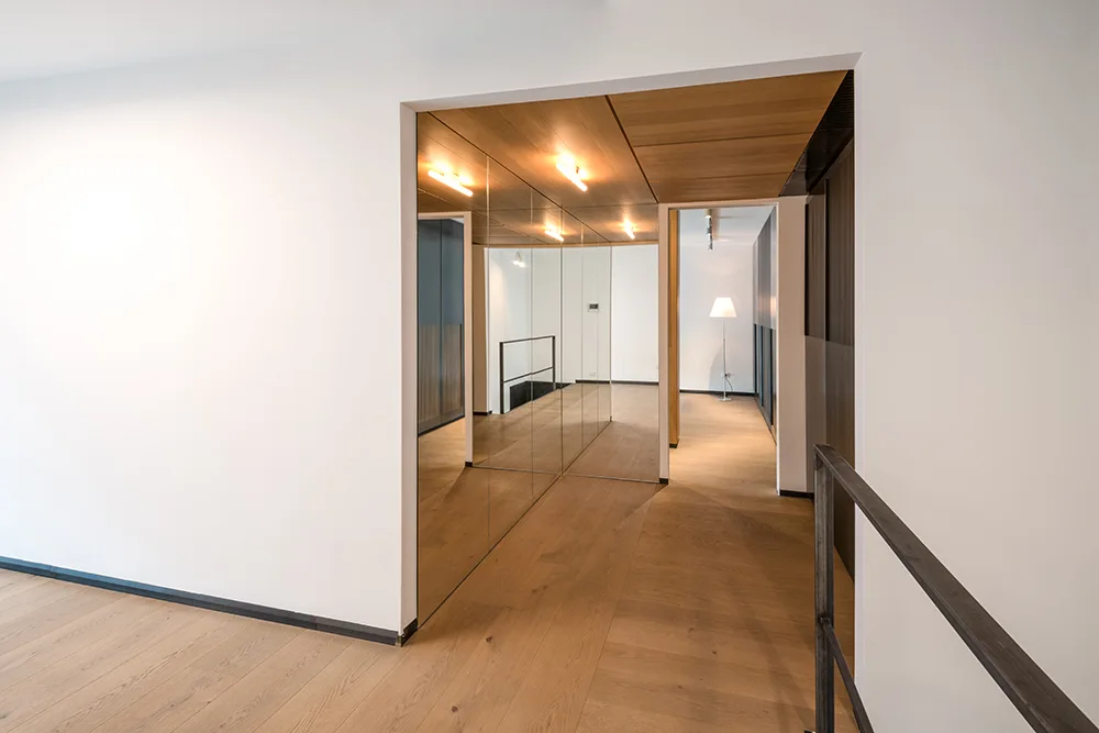 A modern hallway features light-toned wood flooring, a large mirrored wall, a wood-paneled ceiling, and a dark metal stair railing.