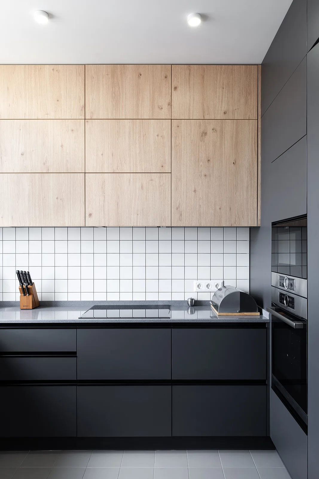 A minimalist kitchen displays a white grid tiled backsplash, light woodgrain upper cabinetry, and dark grey base cabinets.