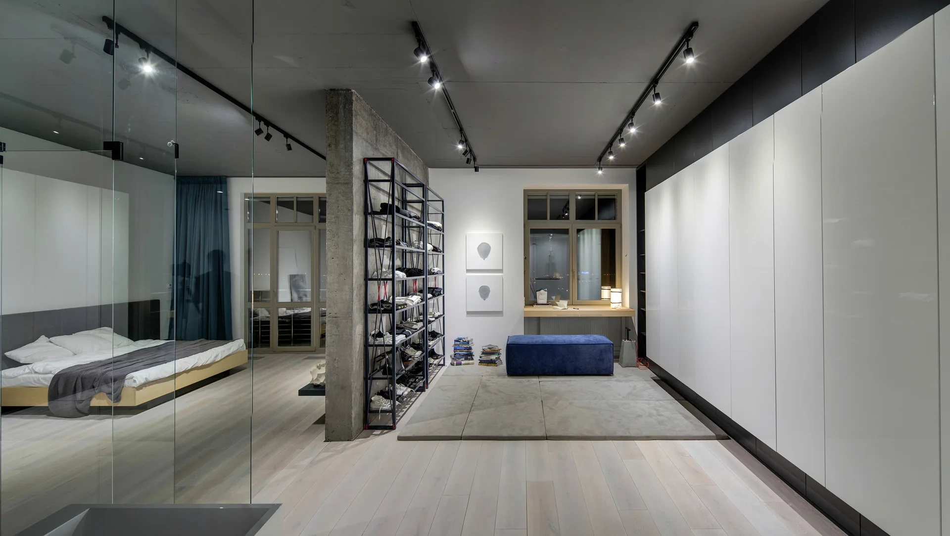 An industrial-style bedroom features a concrete partition wall next to an open metal shelf, a blue bench, large white glossy wardrobes, and a platform bed.