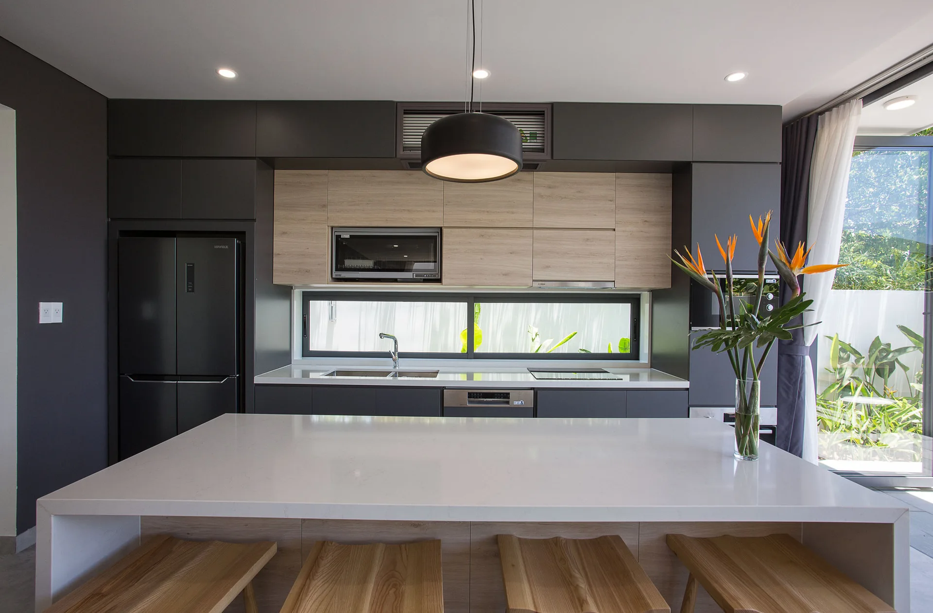 A modern kitchen with dark charcoal cabinetry, light wood upper cabinets, a white island with stools, and a long horizontal backsplash window.