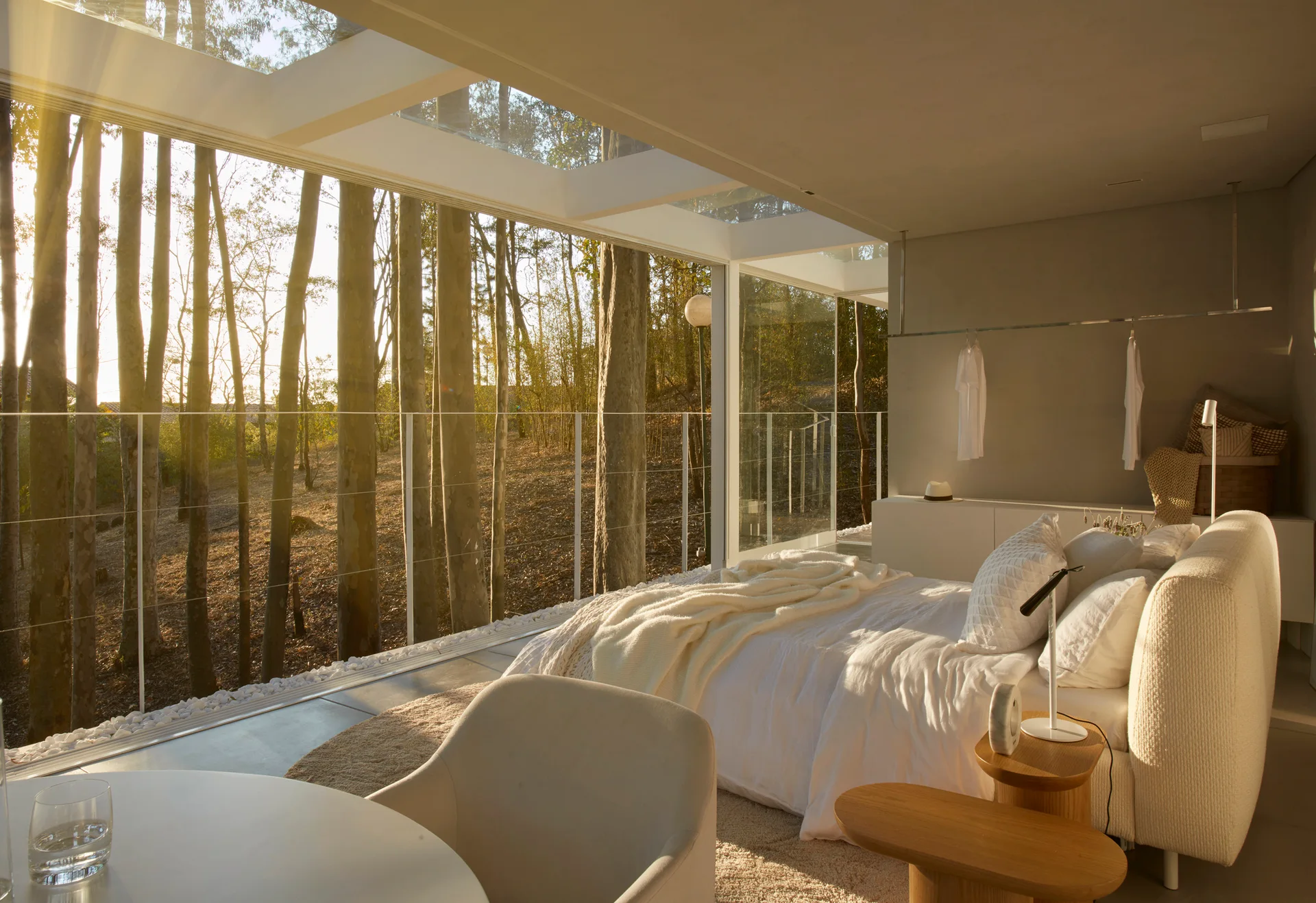 A minimalist bedroom featuring a white upholstered bed, wooden side tables, and floor-to-ceiling glass walls that look out onto a dense forest at sunset.