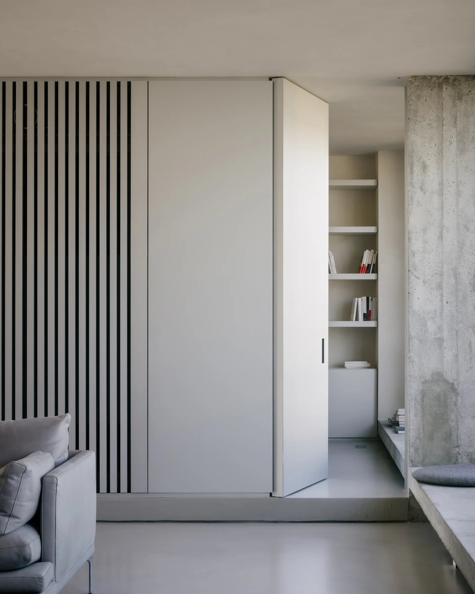 A minimalist white cabinet door opens to reveal a built-in bookshelf next to a slatted wall panel and an exposed concrete pillar.