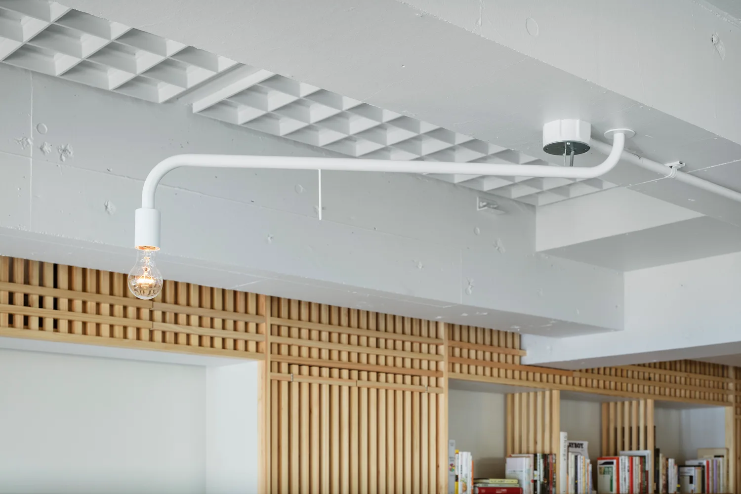 A minimalist white metal tubular light fixture is mounted on a painted concrete waffle ceiling above light wood slatted shelving.
