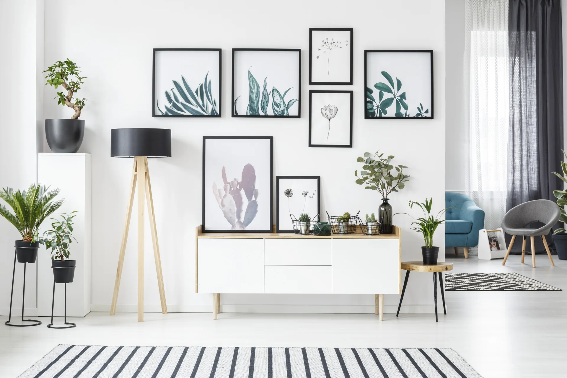 A white sideboard sits below a botanical gallery wall, paired with a wooden tripod floor lamp and a black and white striped rug.