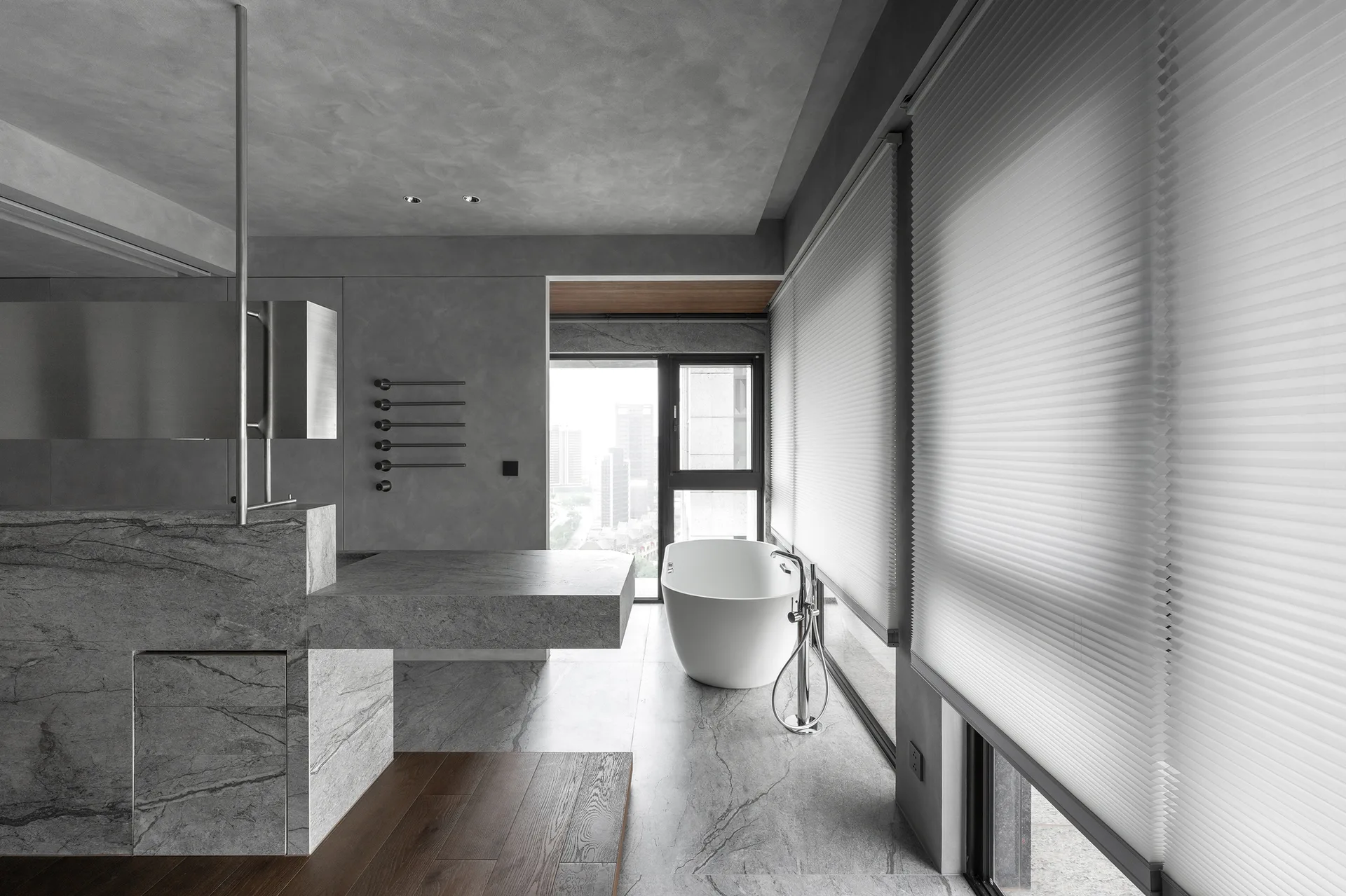 A minimalist grey bathroom featuring a modern freestanding bathtub, a massive stone block vanity, and plaster walls next to large windows with pleated blinds.