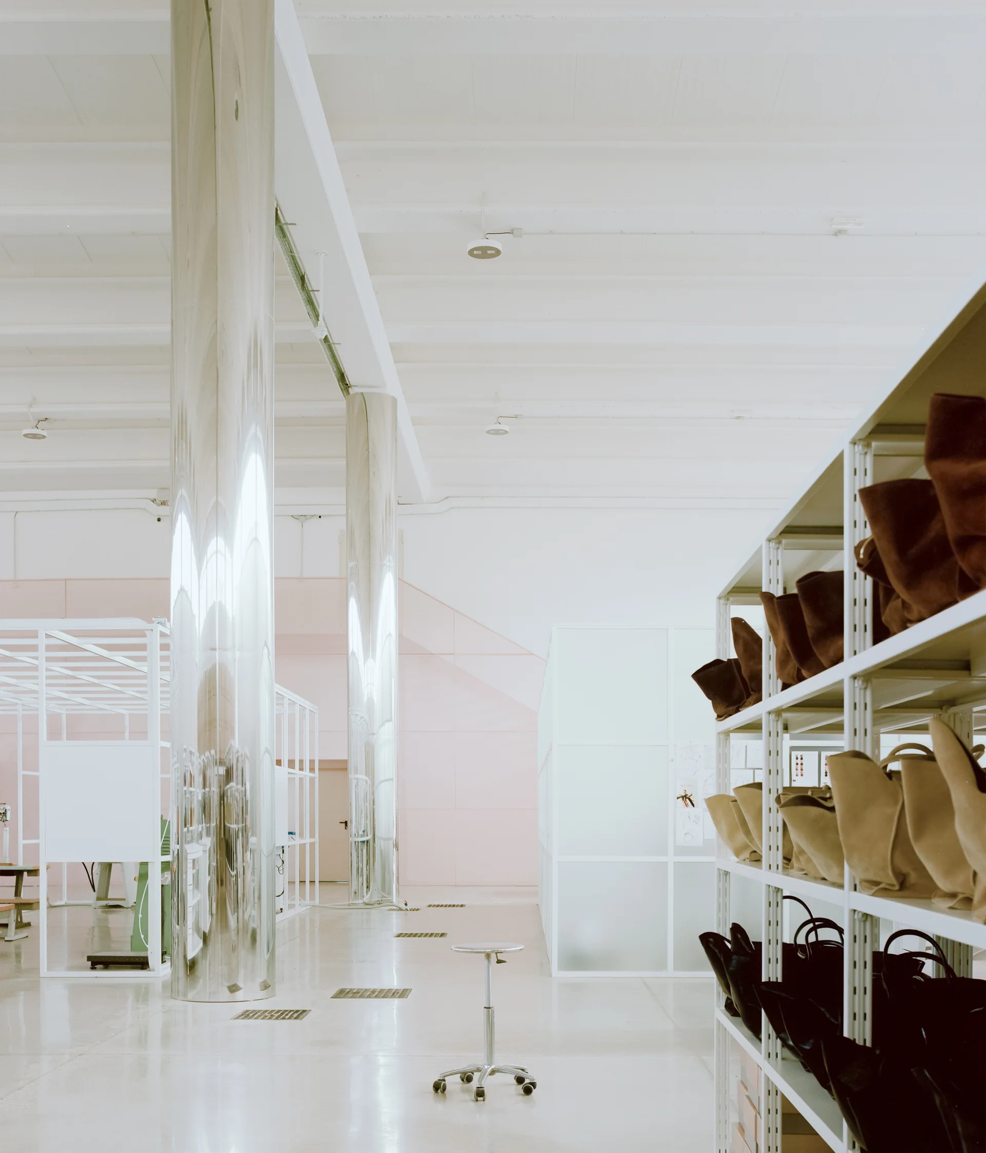 A bright industrial studio with tall reflective chrome columns, open white shelving holding leather bags, and a glossy light concrete floor.