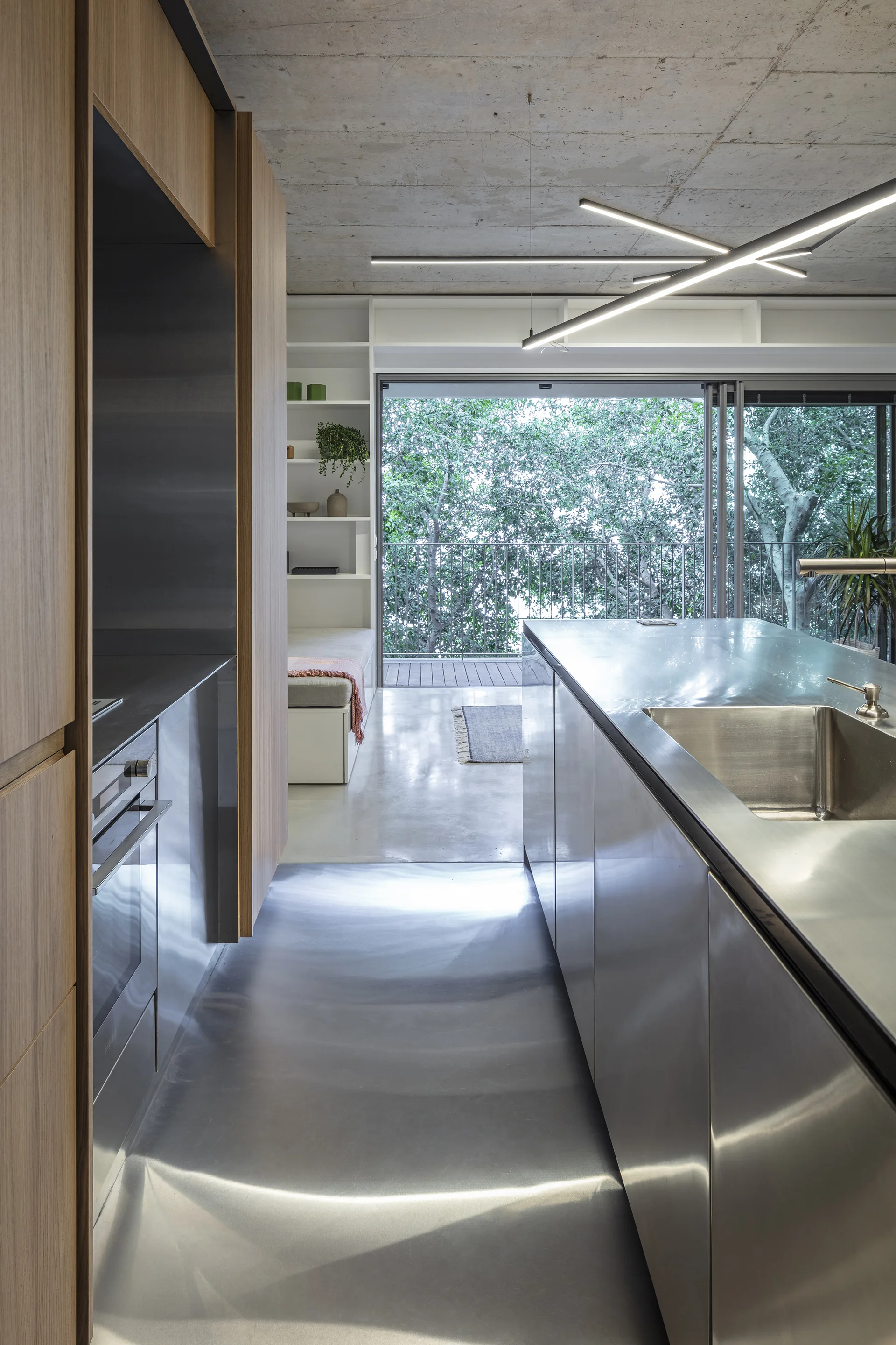 A modern kitchen featuring a stainless steel island and wood cabinets beneath an exposed concrete ceiling with sliding glass doors opening to trees.