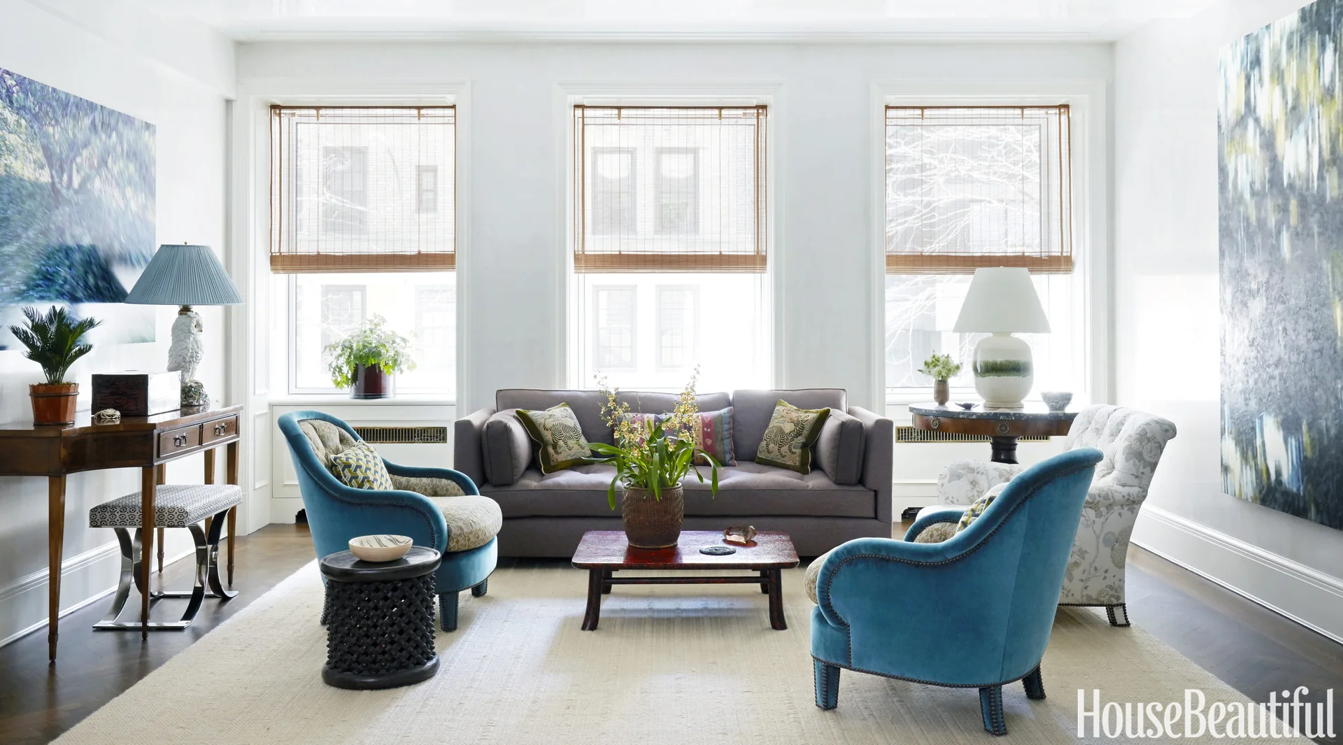 A bright living room features a grey sofa flanked by two blue velvet armchairs with three windows wearing woven shades behind them.