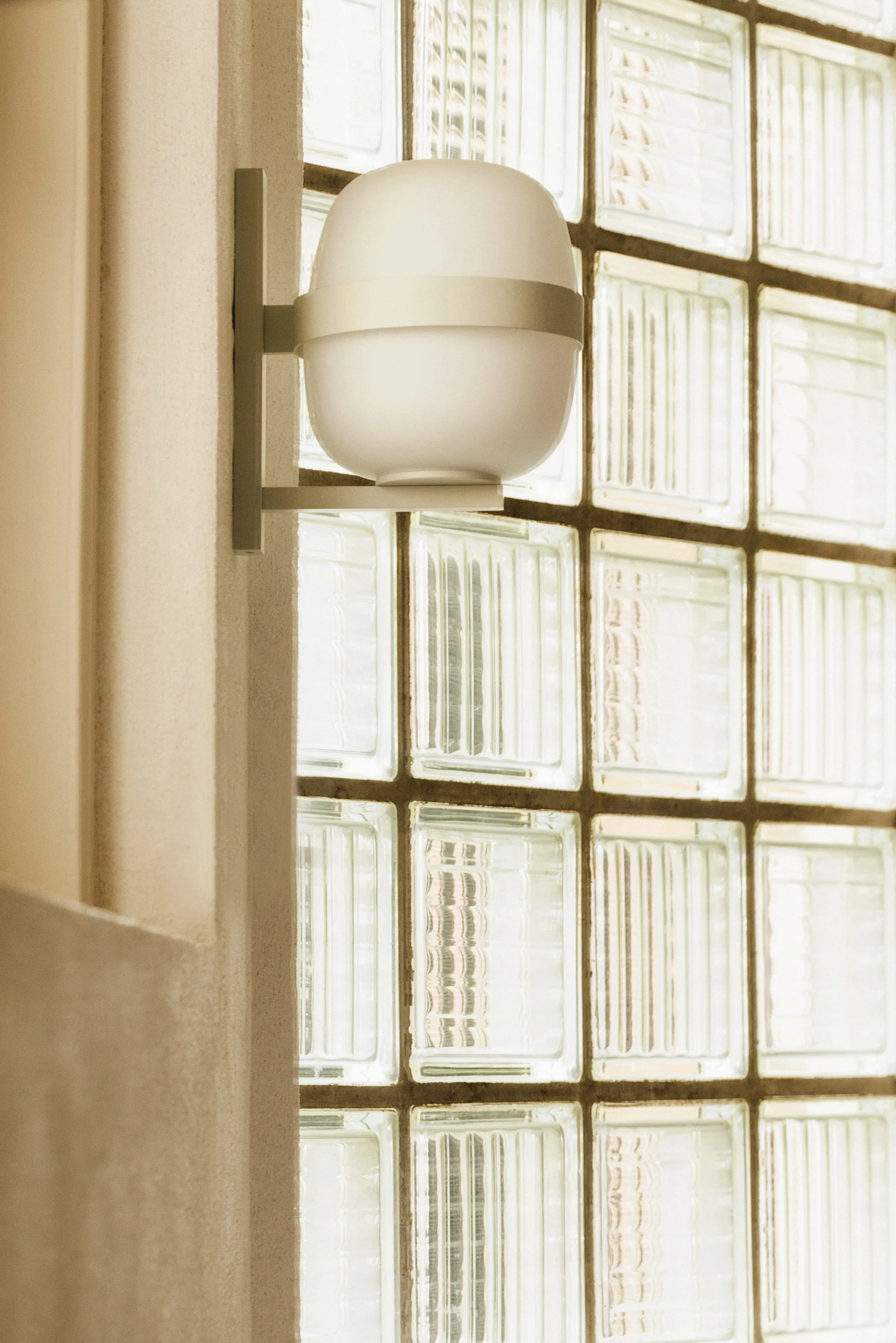 A close-up of a matte white rounded wall sconce mounted on a beige column beside a grid-patterned glass block window.