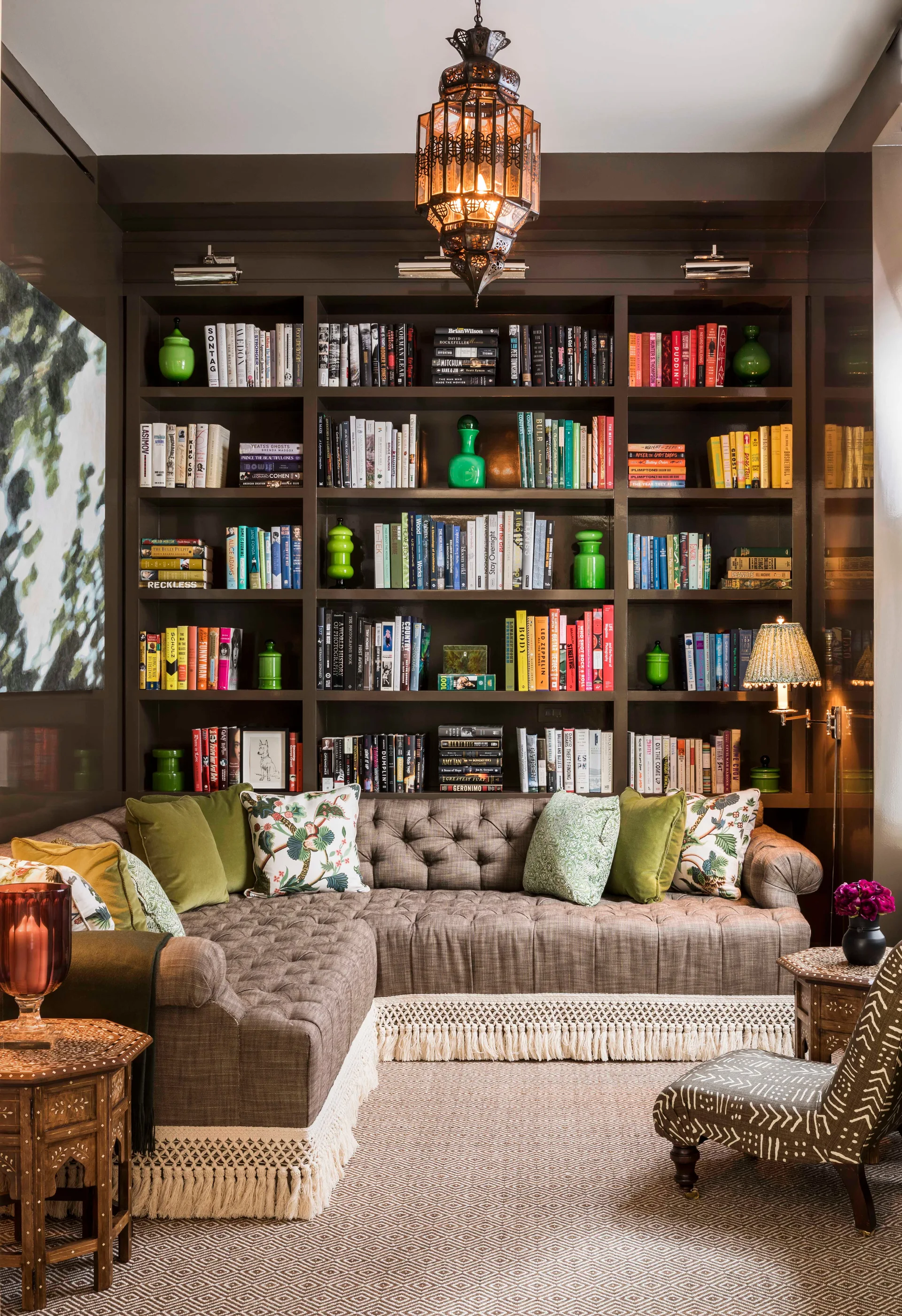 A cozy library features a high-gloss dark brown built-in bookcase behind an L-shaped sectional sofa decorated with a white tassel fringe.