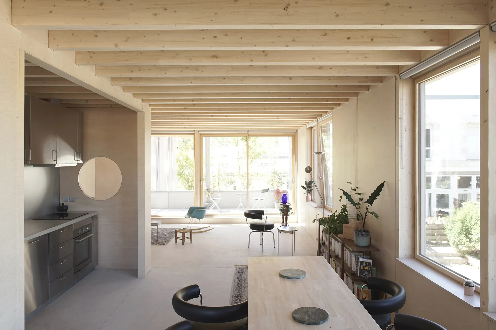 A minimalist open-plan dining and kitchen space featuring light plywood walls, exposed wooden ceiling beams, and stainless steel cabinetry.