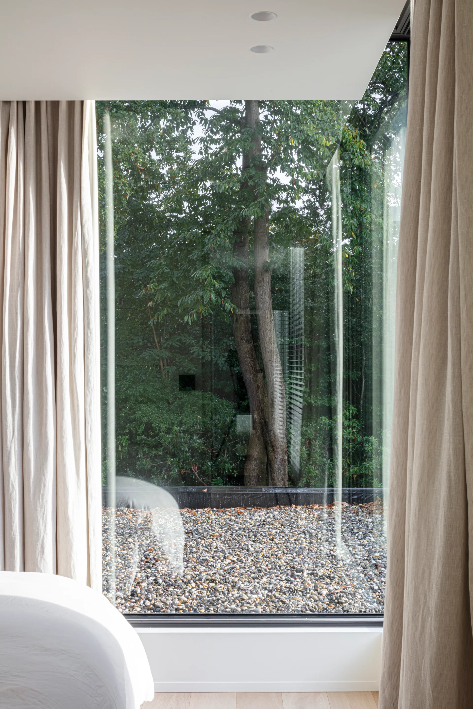 A floor-to-ceiling window in a minimalist bedroom is flanked by beige linen curtains and opens to a pebble-lined garden with trees.