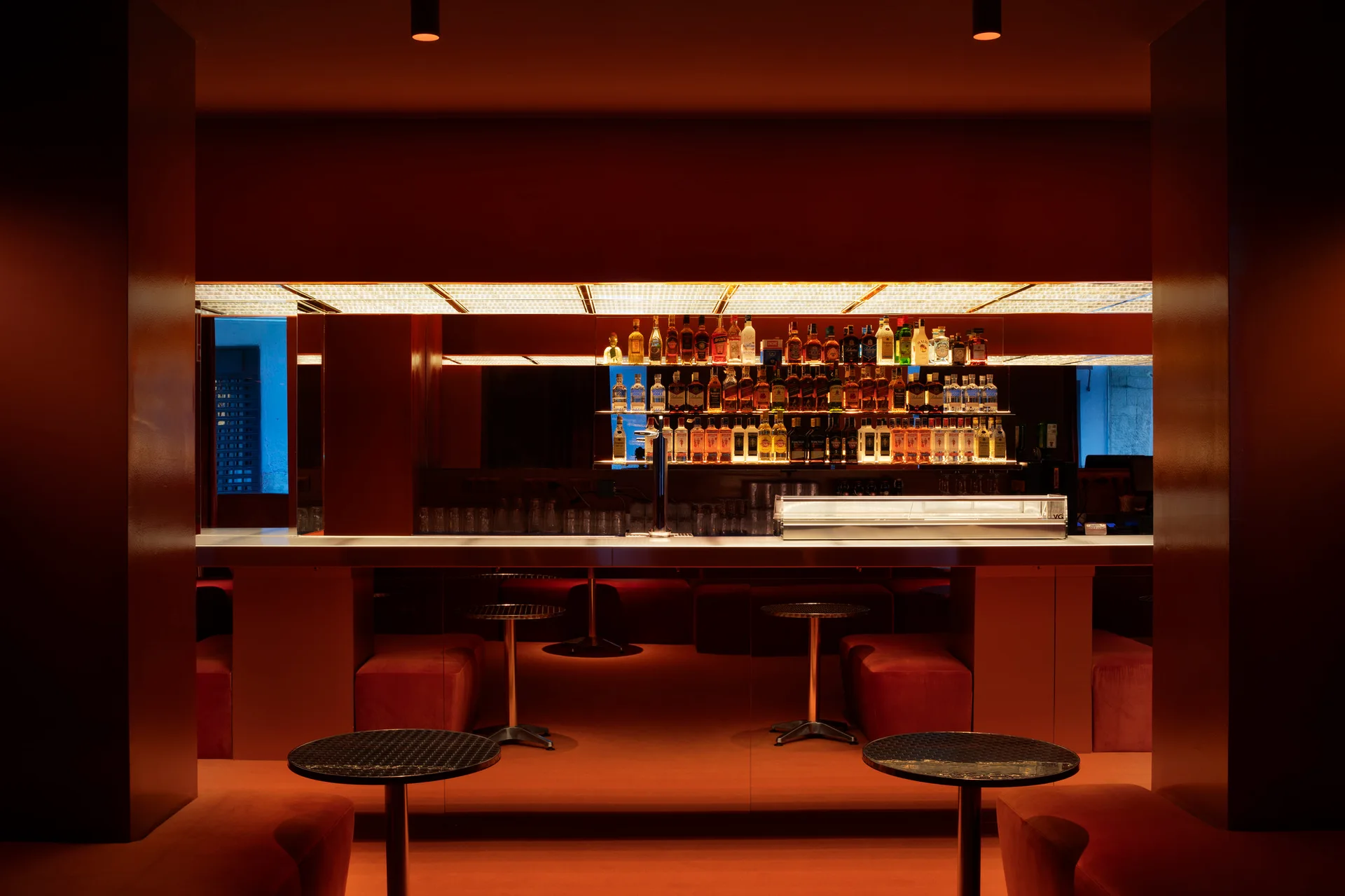 A dimly lit bar interior saturated in deep red paint features a long counter, velvet-upholstered seating, and backlit glass liquor shelving.