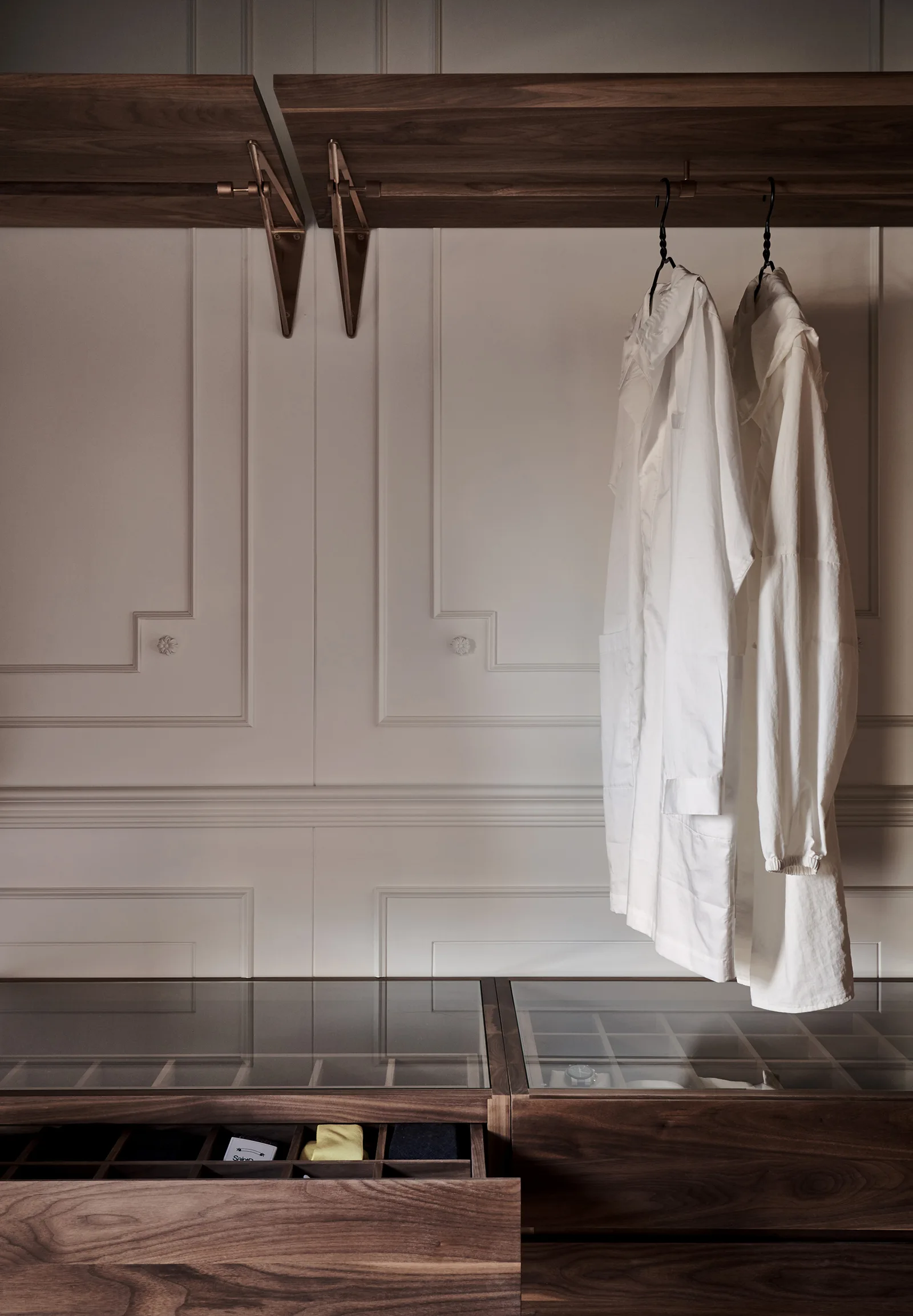 A dark wood walk-in closet system features hanging garments, a metal rod, and glass-topped dresser drawers against a beige paneled wall.