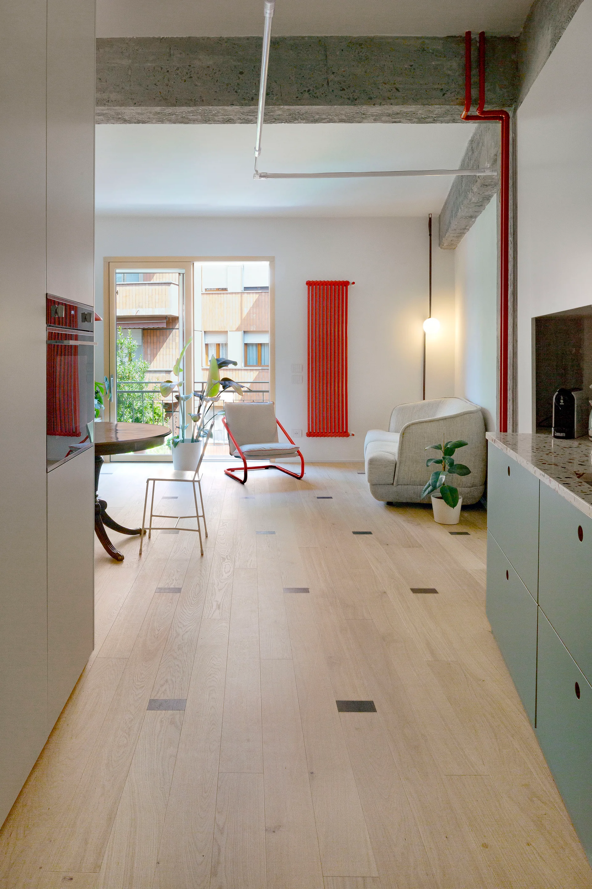 An open-plan living room features light wood floors with dark inlays, a grey sofa, a red vertical radiator, and exposed concrete ceiling beams.