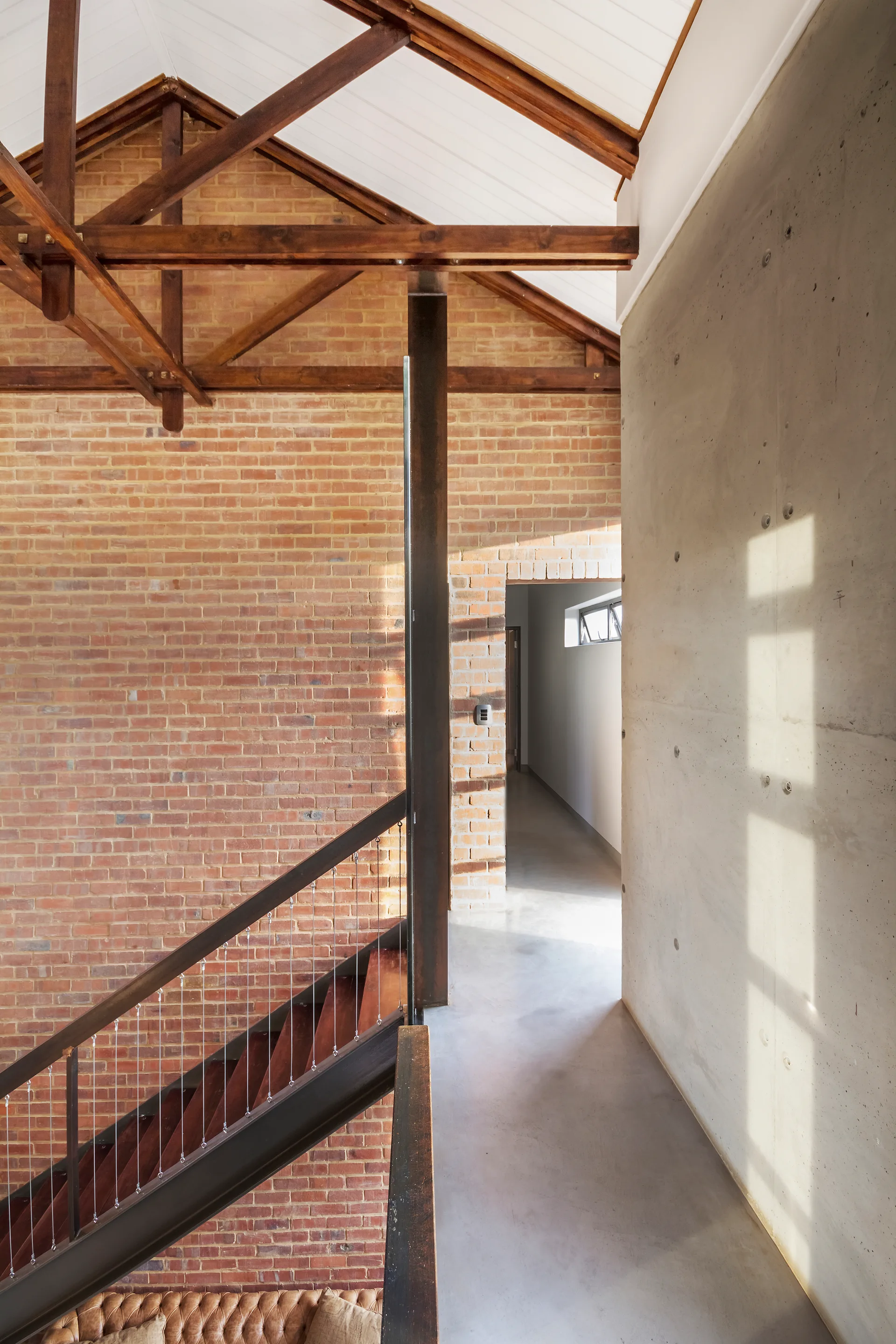 An industrial hallway featuring a wood staircase with cable railing, an exposed brick wall, and a smooth concrete wall under timber ceiling trusses.