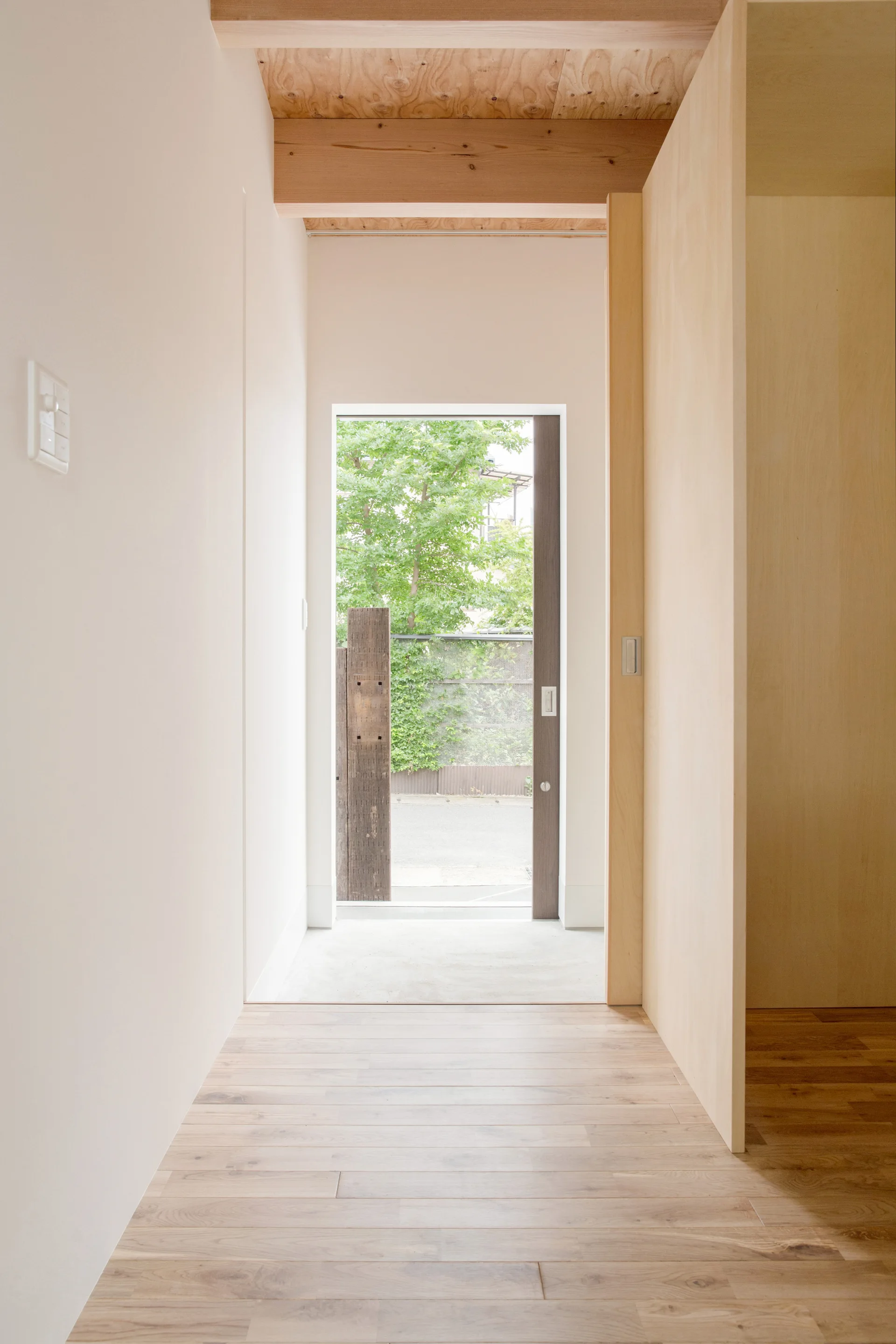 A minimalist entrance hallway features light wood flooring, a pale plywood partition, and exposed ceiling beams leading to an exterior glass door.