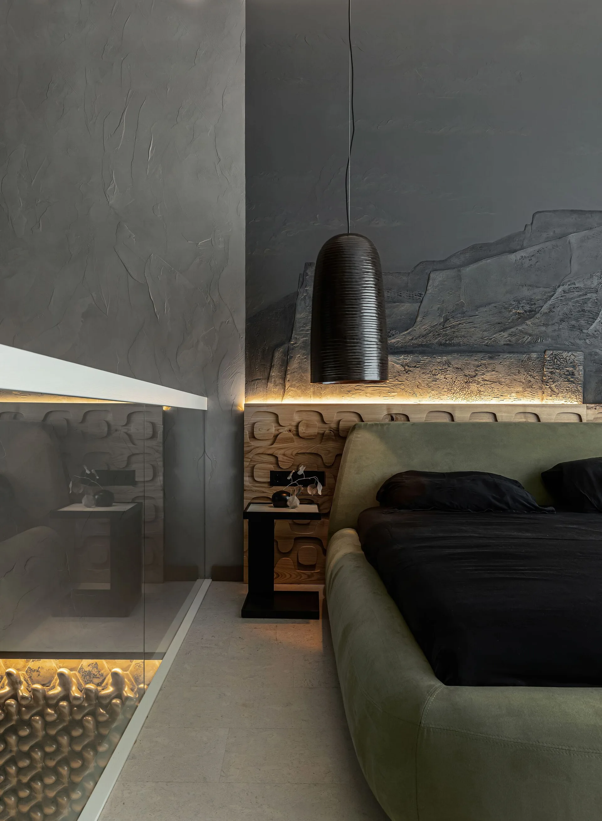 A modern bedroom with a green upholstered bed, a textured gray plaster wall, a carved wood headboard panel, and warm integrated backlighting.