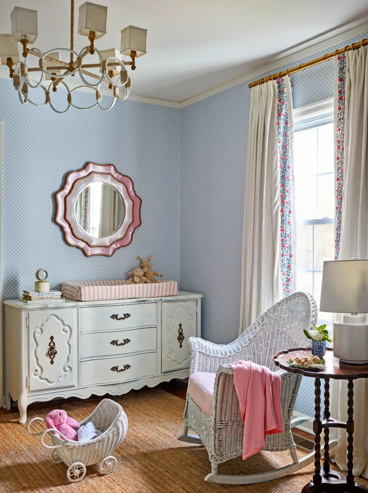A traditional nursery features blue patterned wallpaper, a white dresser with a changing pad, a pink scalloped mirror, and a white wicker rocking chair.